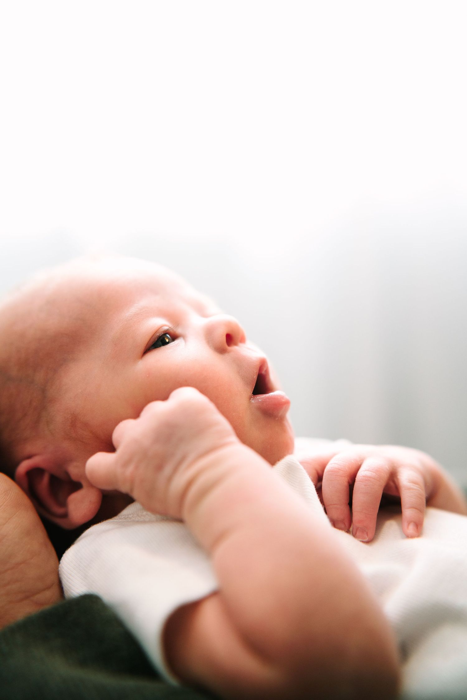 Close-up of a newborn baby with mouth open and tiny hand raised, gazing upward in soft natural light.