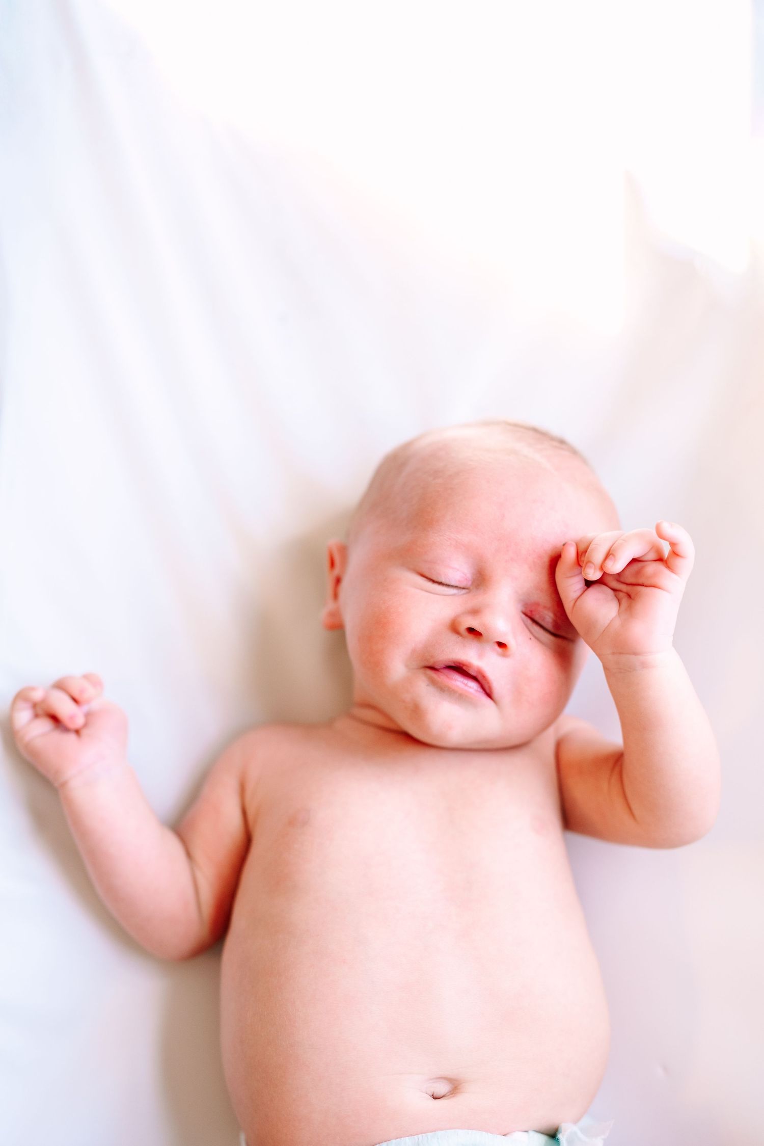 Newborn baby lying on white blanket with eyes closed, rubbing eye with tiny fist in peaceful sleep.