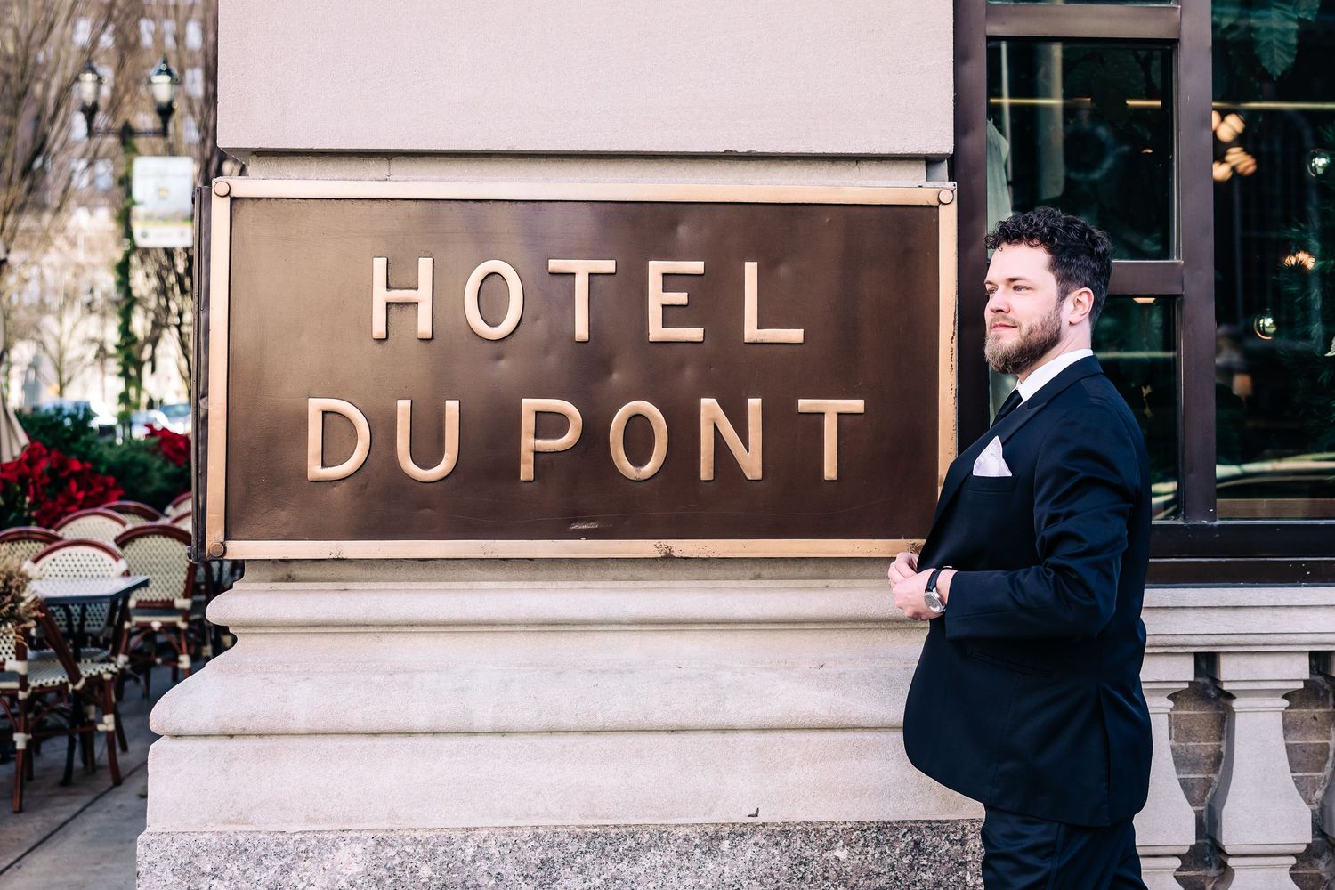 A smiling man in a suit poses beside the Hotel Du Pont sign on an elegant stone building exterior Hotel DuPont wedding
