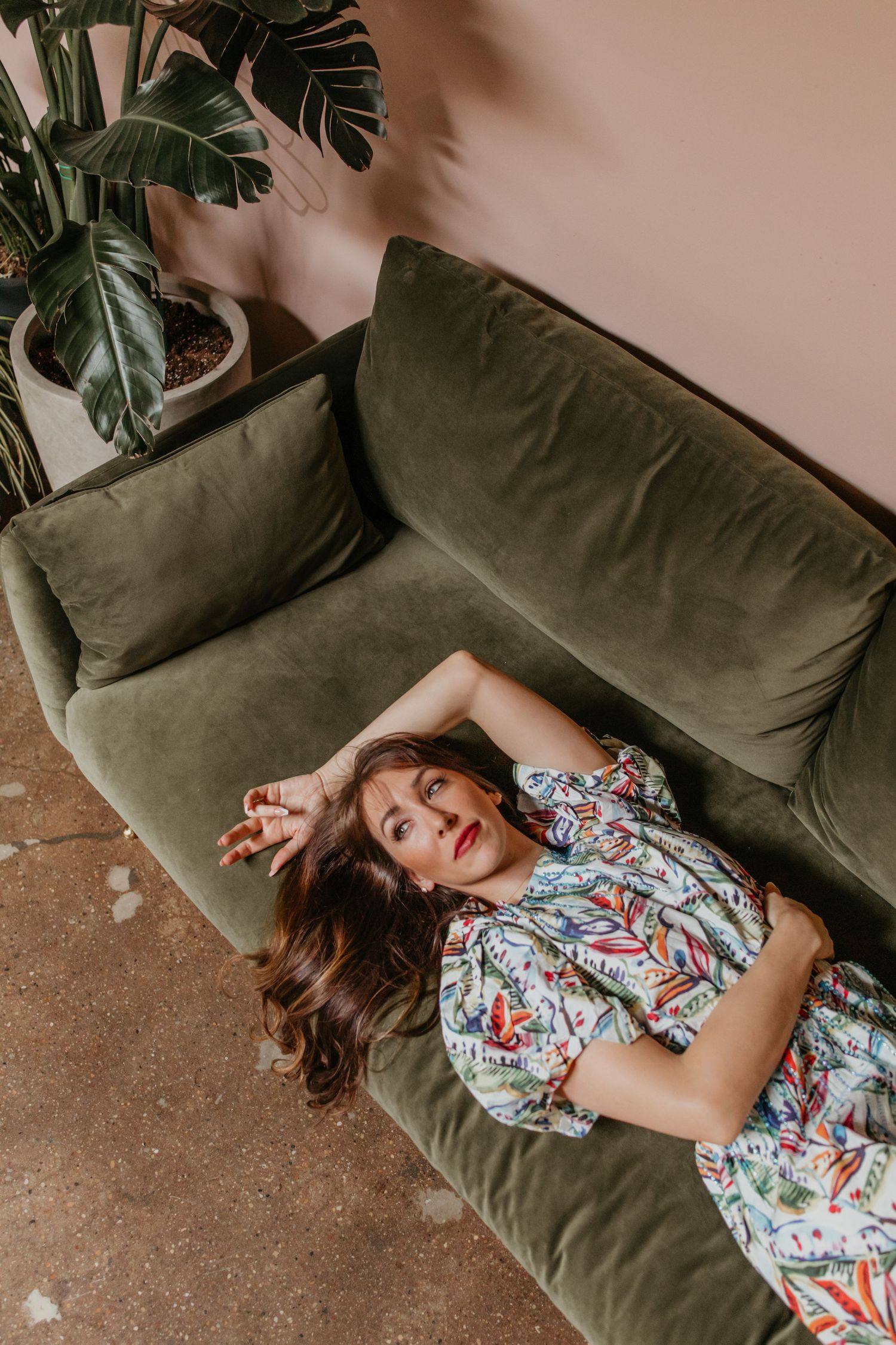 Person in floral dress lounging dramatically on olive green velvet couch with plants.