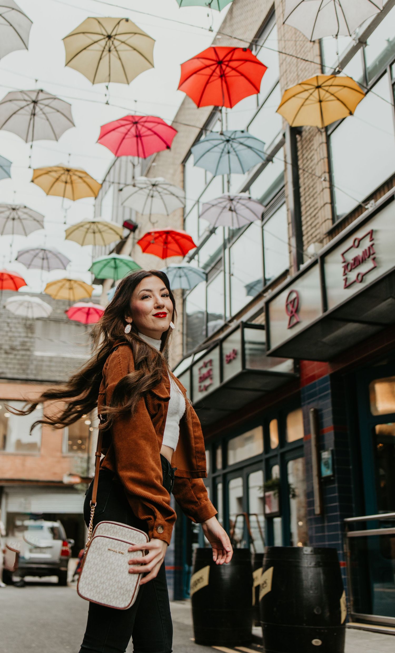 Colorful umbrellas hang from ceiling over person in brown jacket walking through urban corridor.