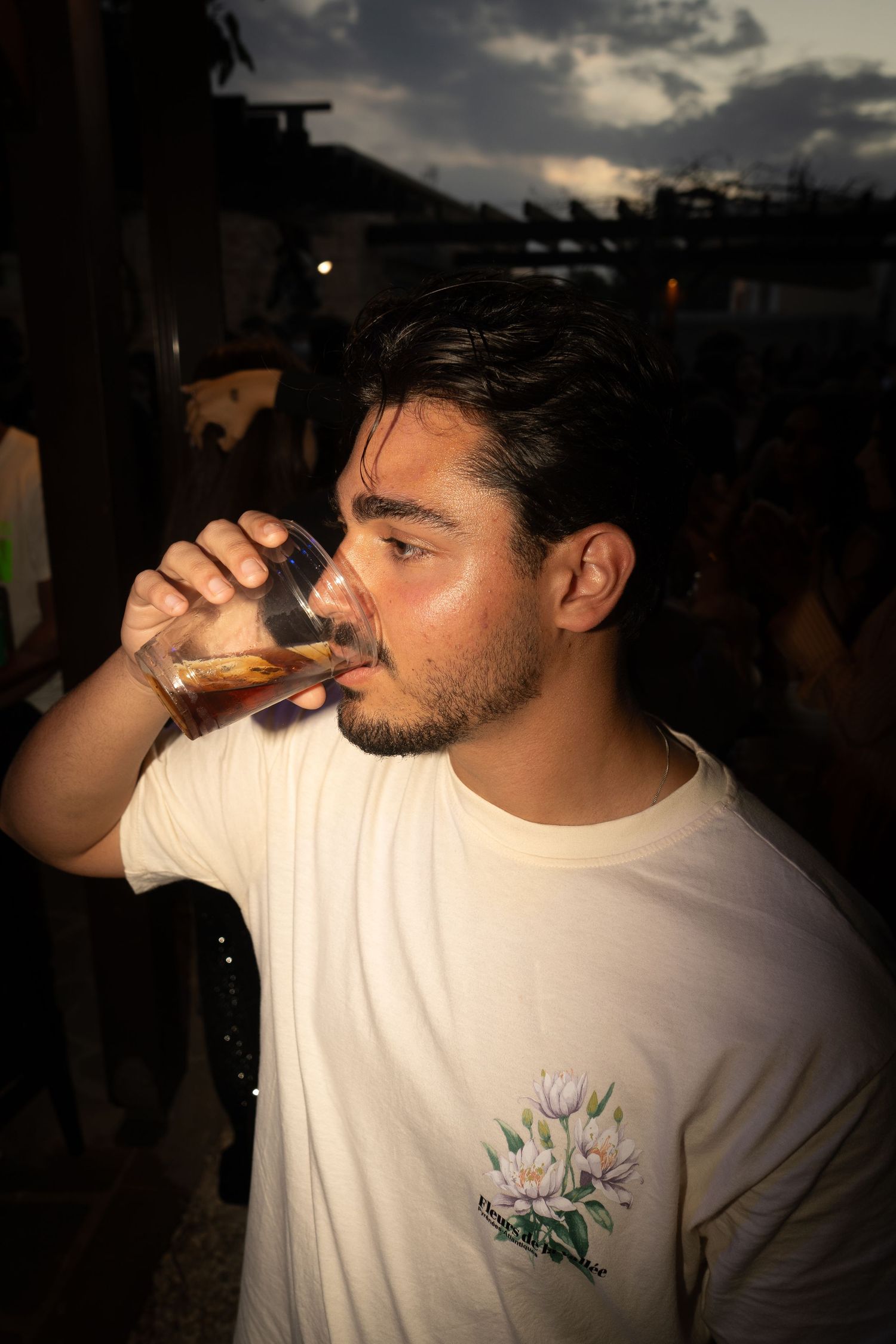 Young man drinking from glass at outdoor evening party in white floral t-shirt.

event, portrait