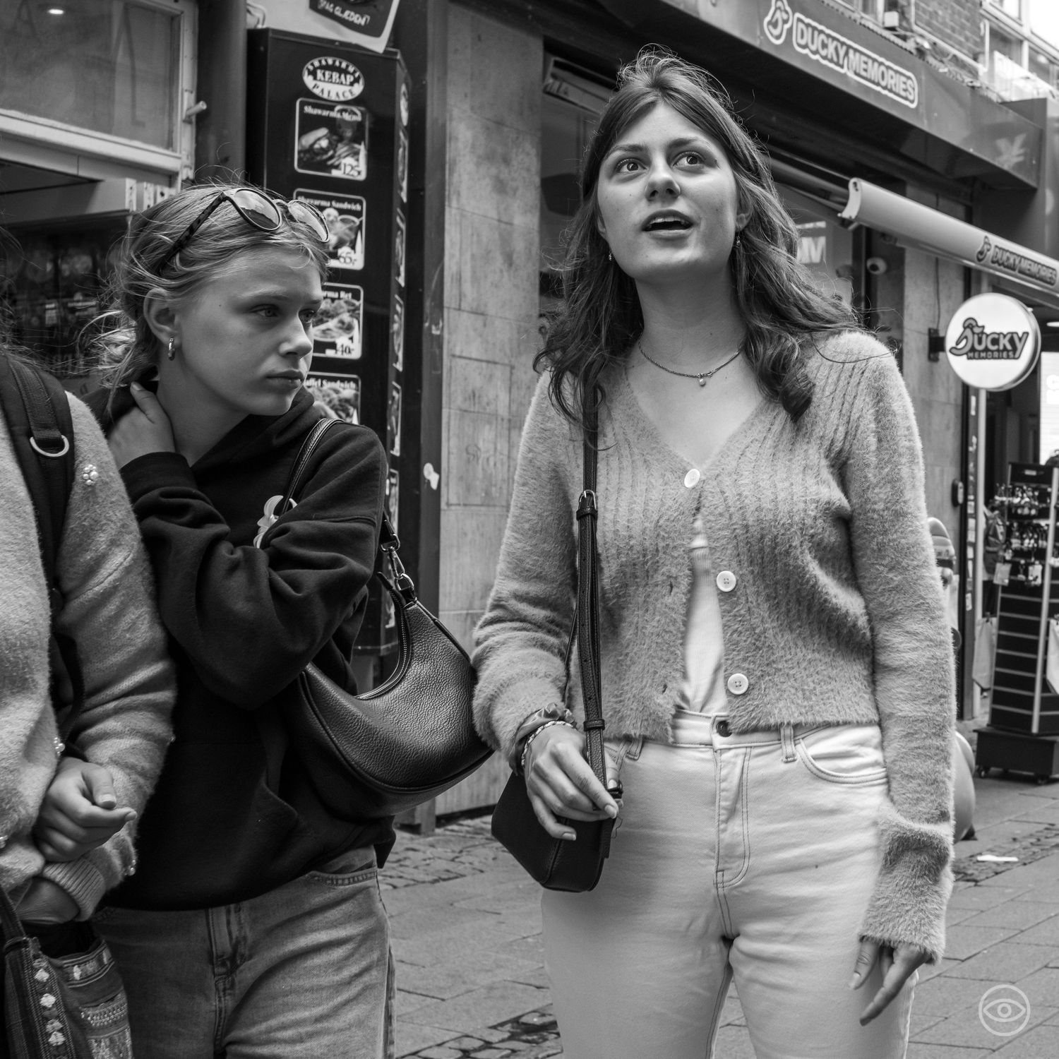 Two young women walking past storefronts, one in a knit cardigan holding a phone, candid black and white street photography.