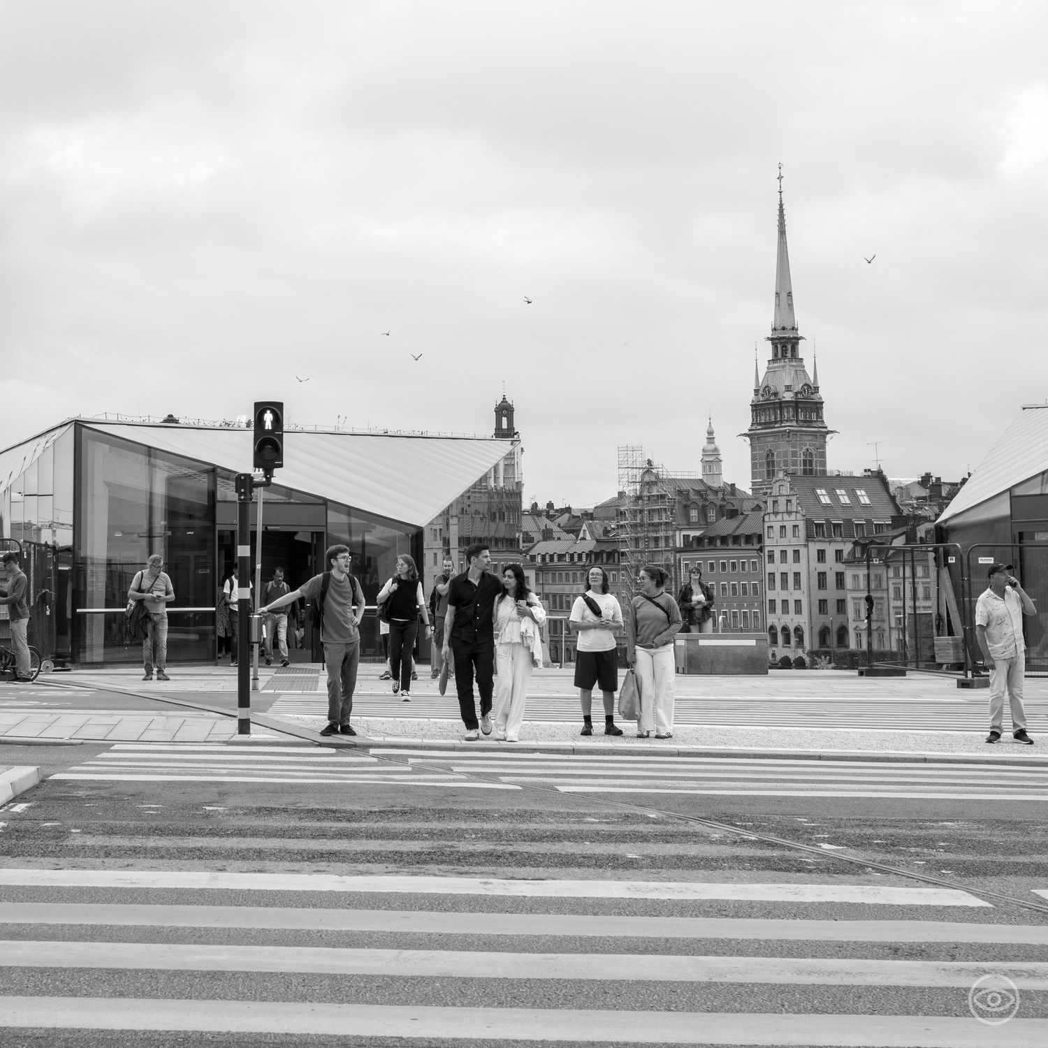 People cross a zebra crossing in Stockholm with a historic church spire visible in the background.