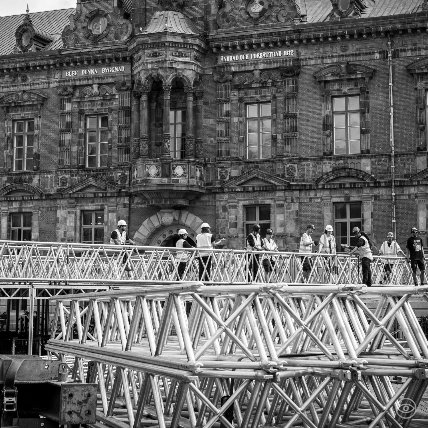 Construction workers in hard hats standing on metal scaffolding truss in front of a historic European building, black and white.