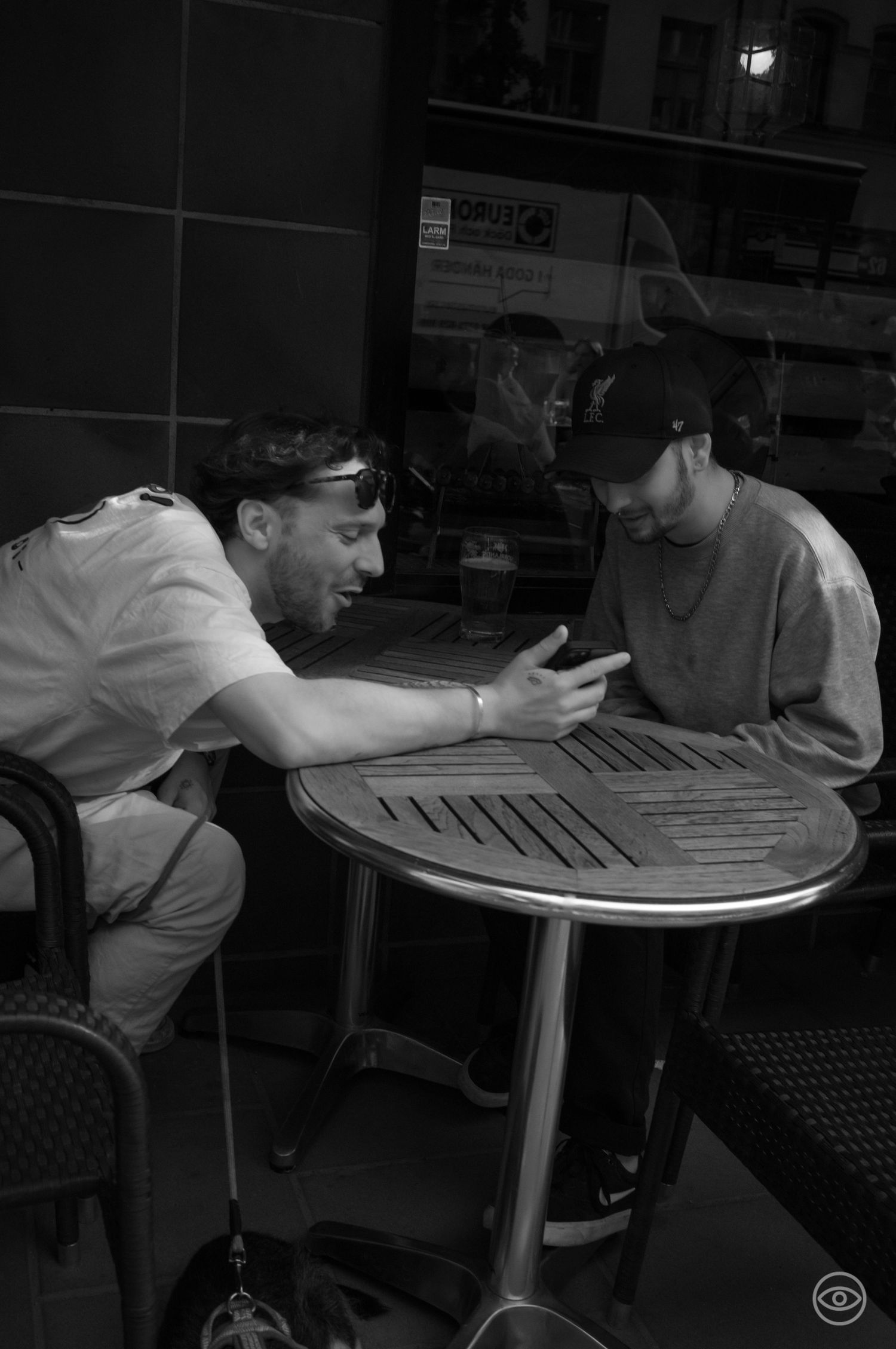 Two men examine a smartphone at a small round bistro table in a black and white street photography shot.