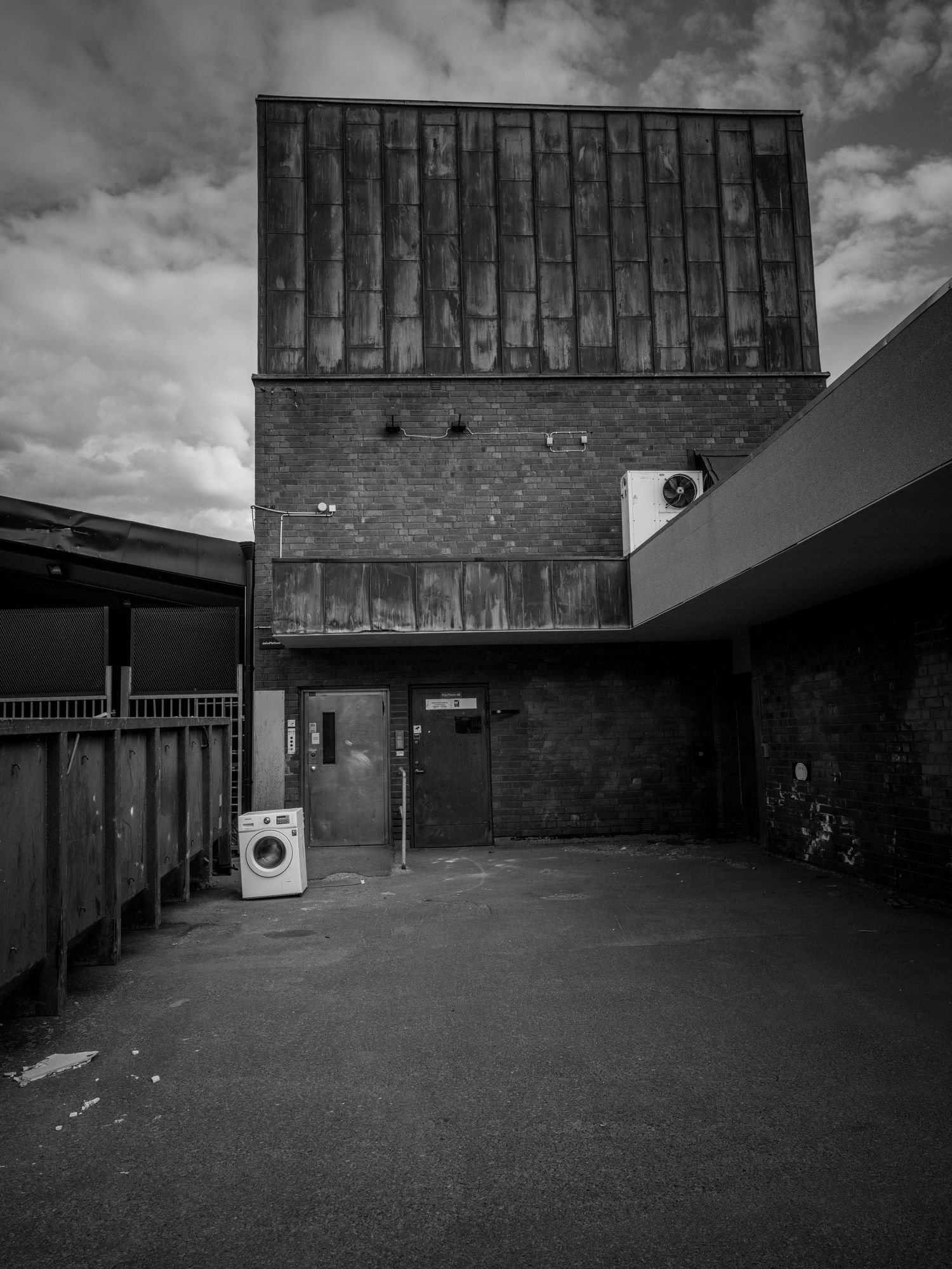 Black and white photo of a brick building with rusty facade and abandoned washing machine outside.