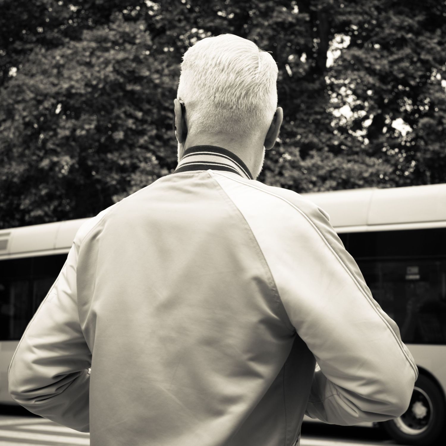 Silver-haired man in striped jacket stands with back to camera near bus, black and white.