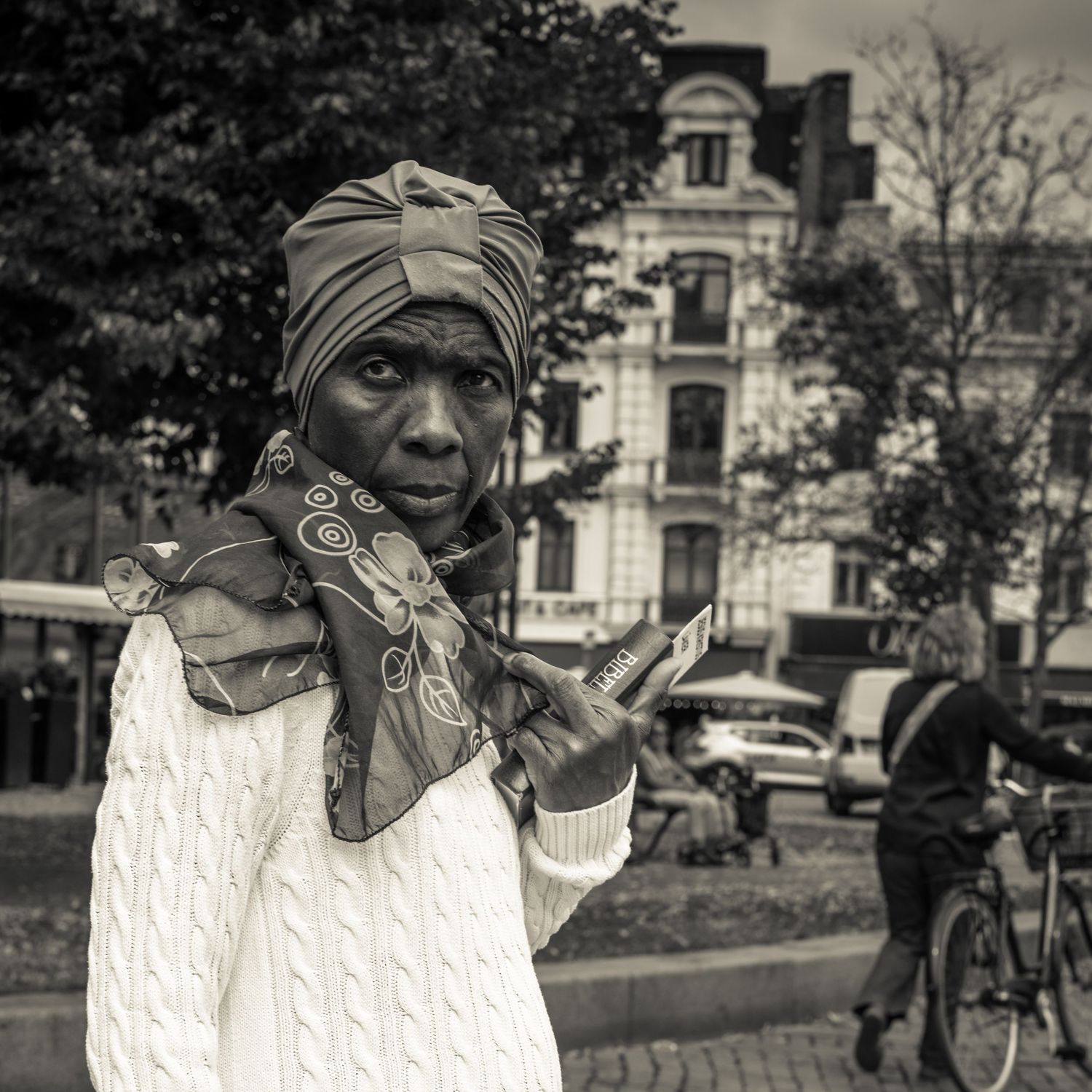 Close-up of African woman in white cable knit sweater and colorful head wrap in city square.