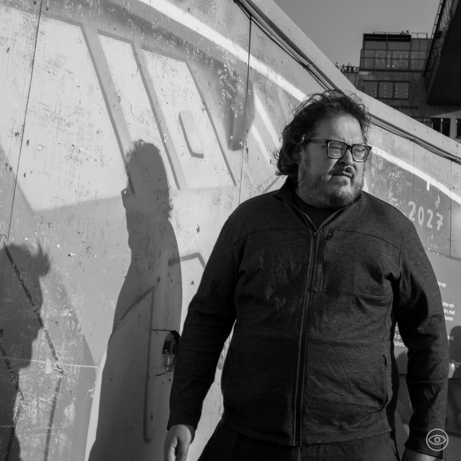 Heavyset man with long hair and glasses standing against a concrete wall in sunlight.