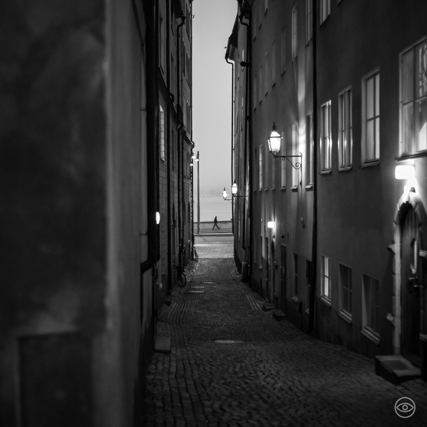 A narrow cobblestone alley lined with old buildings and vintage lanterns leading to open water at dusk, black and white photo.