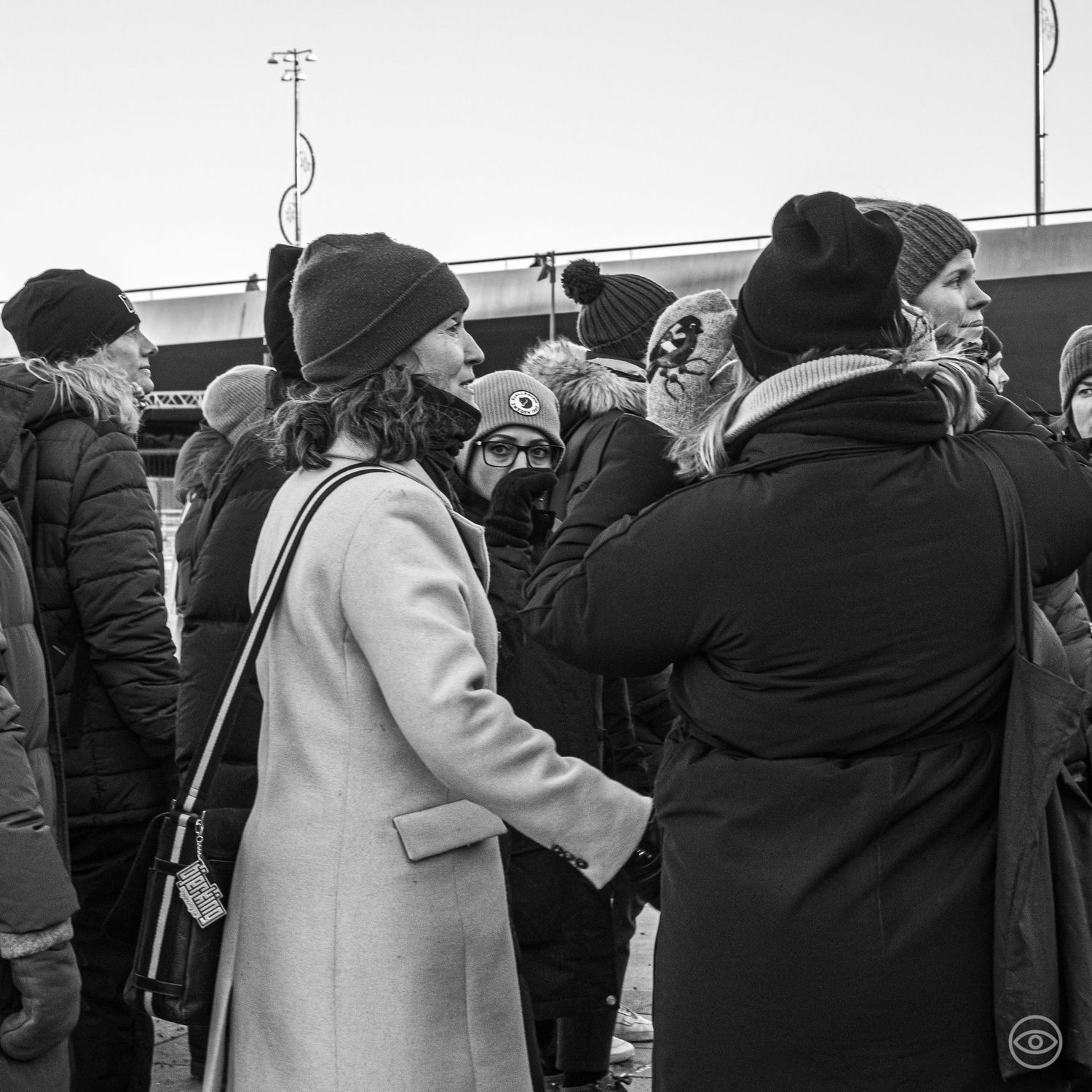 Group of people in winter coats and knit hats gathered outdoors in a black and white street photo.