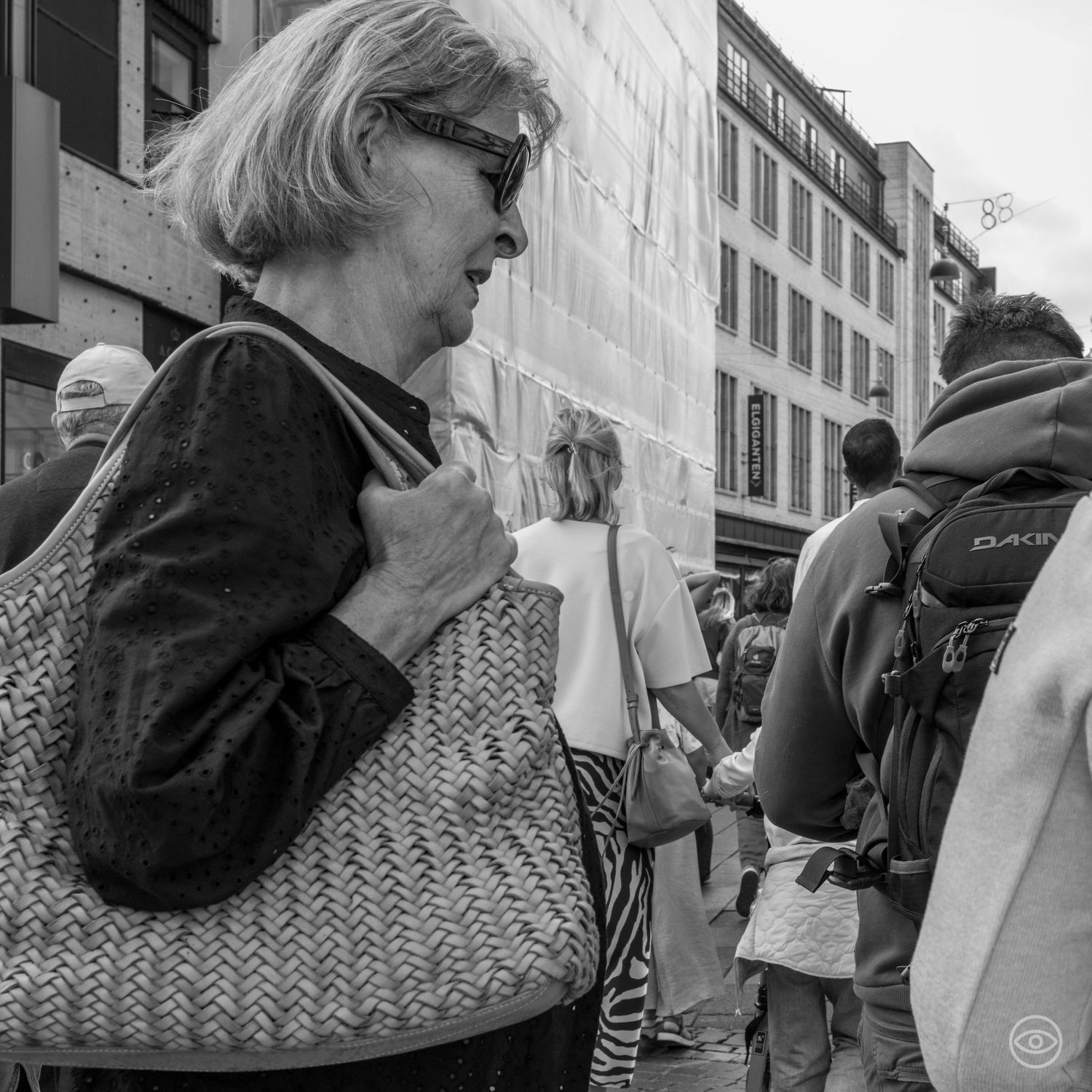 Older blonde woman with sunglasses carrying a woven bag on a busy city street in black and white.