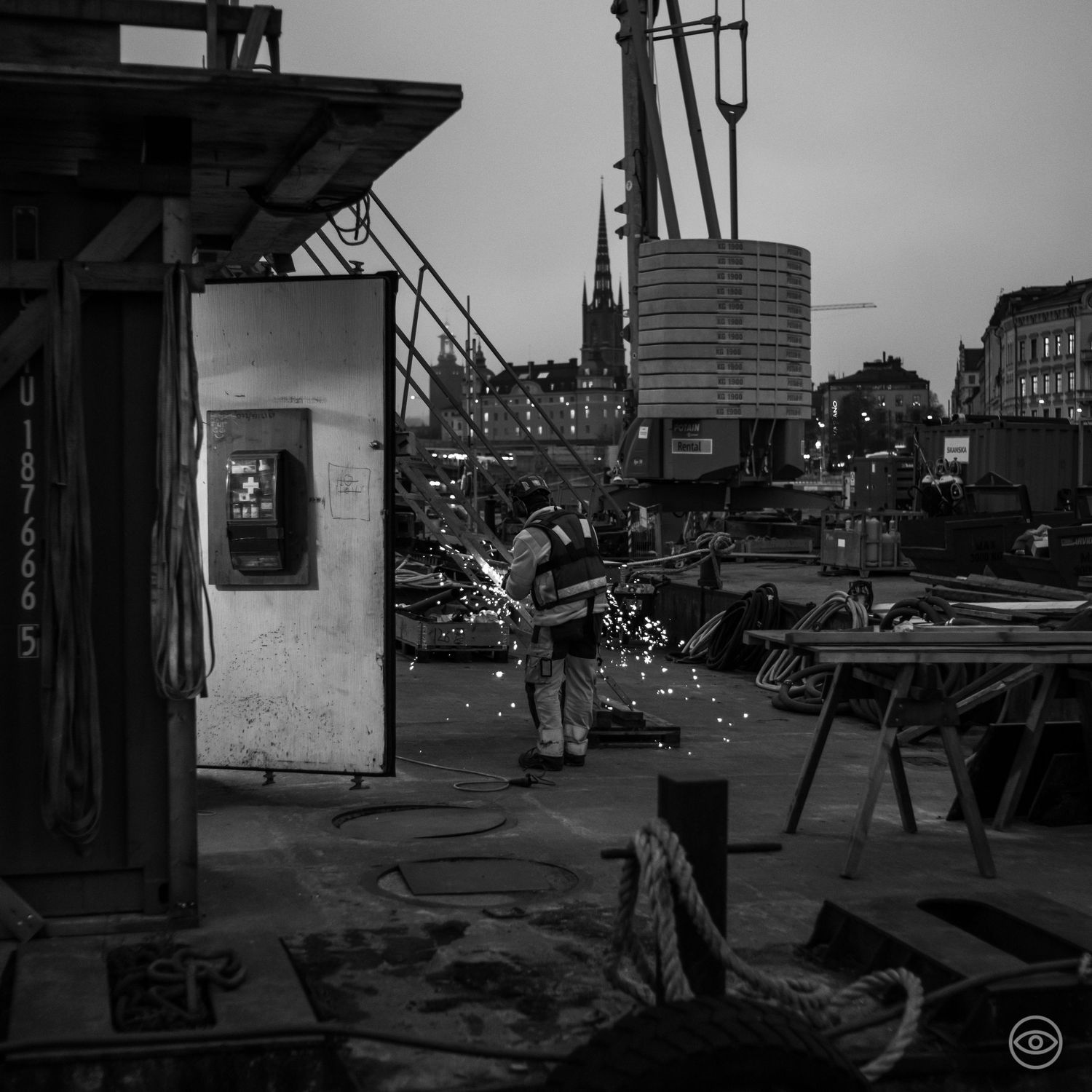 A welder creating sparks while working at a shipyard construction site at dusk with a church spire visible, black and white photo.