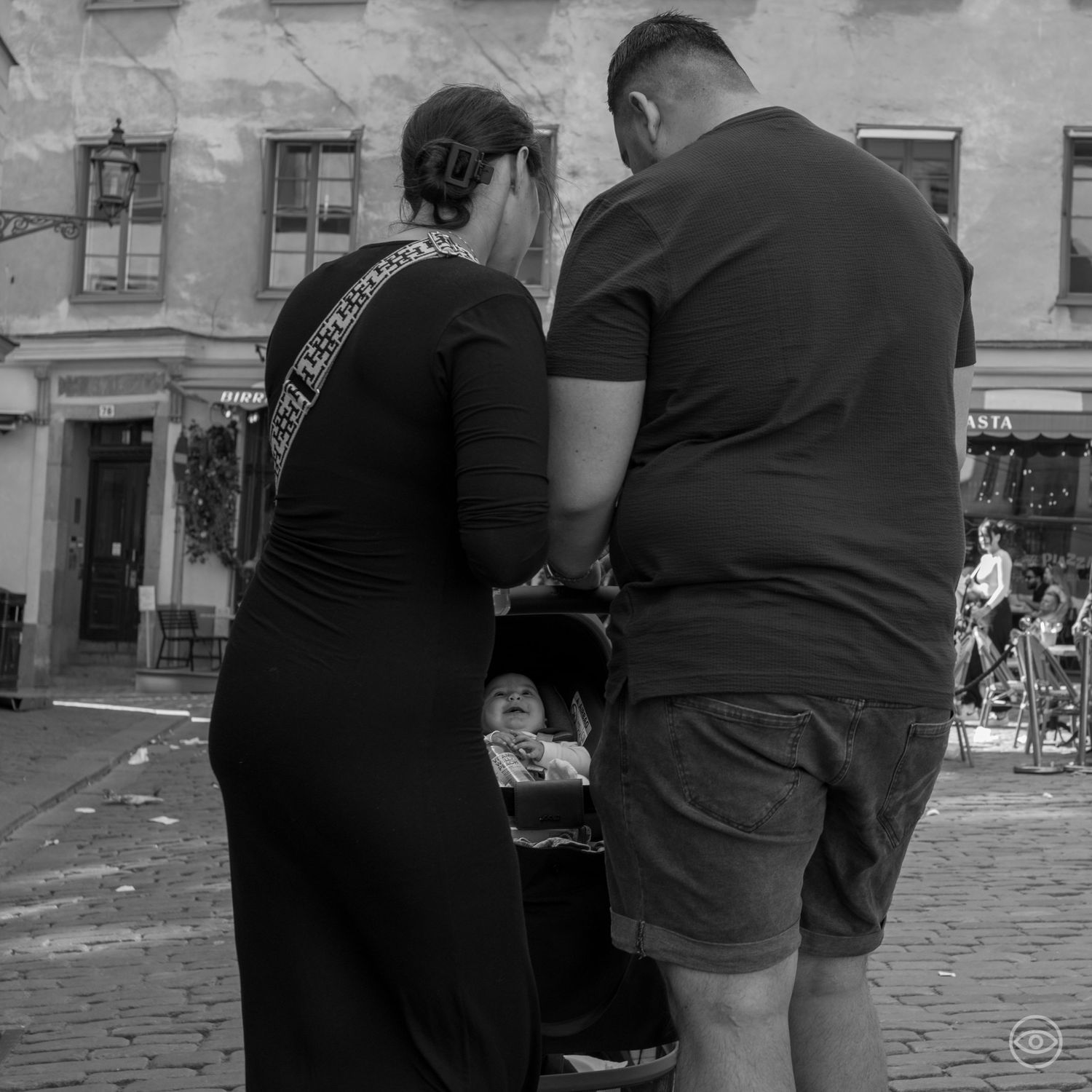 Couple leaning over a baby stroller on a cobblestone street, black and white candid photography.