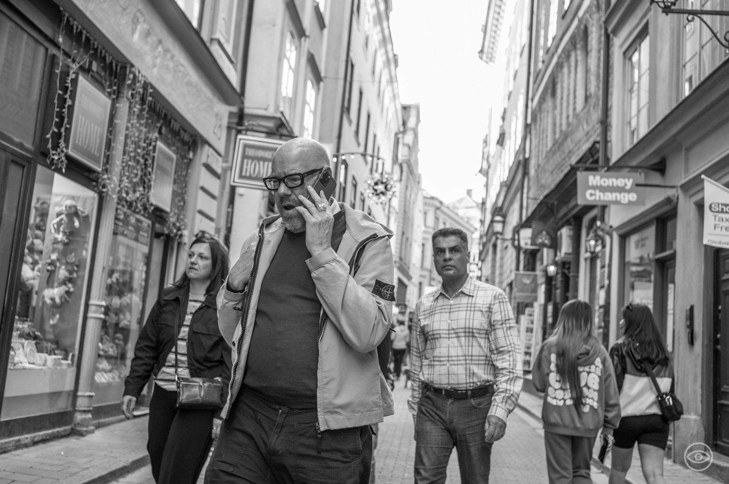 Bald man talking on phone walks through a narrow European shopping street in black and white photography.