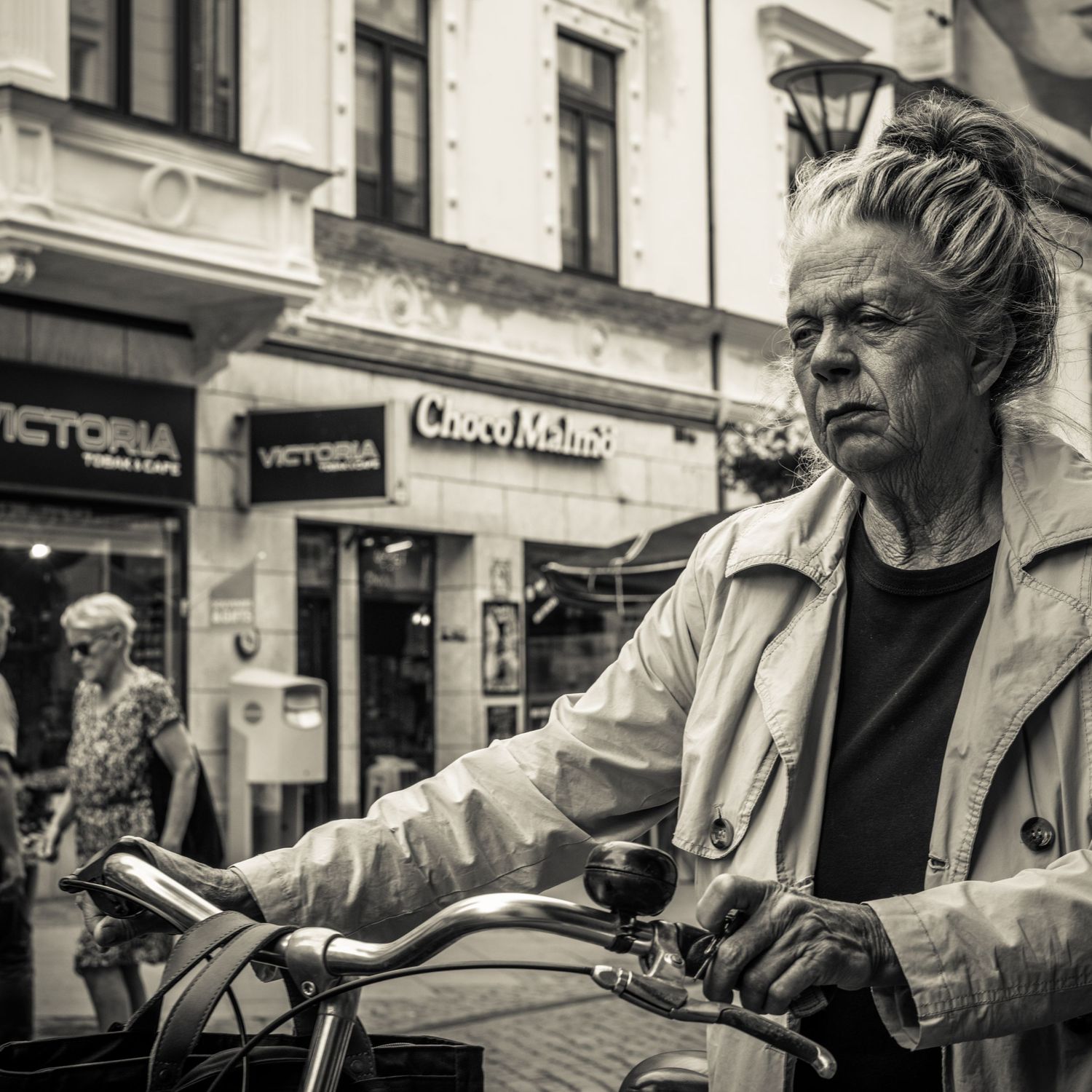 Older woman with upswept hair pushes bicycle past Victoria cafe on urban street, black and white.