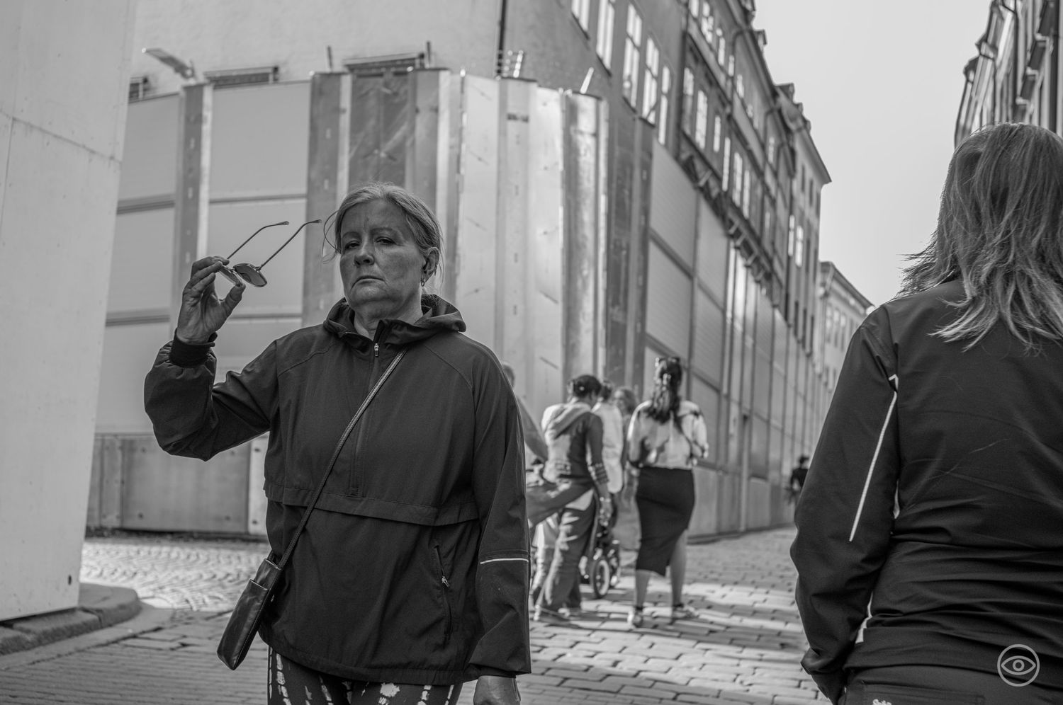 Older woman in a jacket holds glasses while walking on a European cobblestone street in black and white.