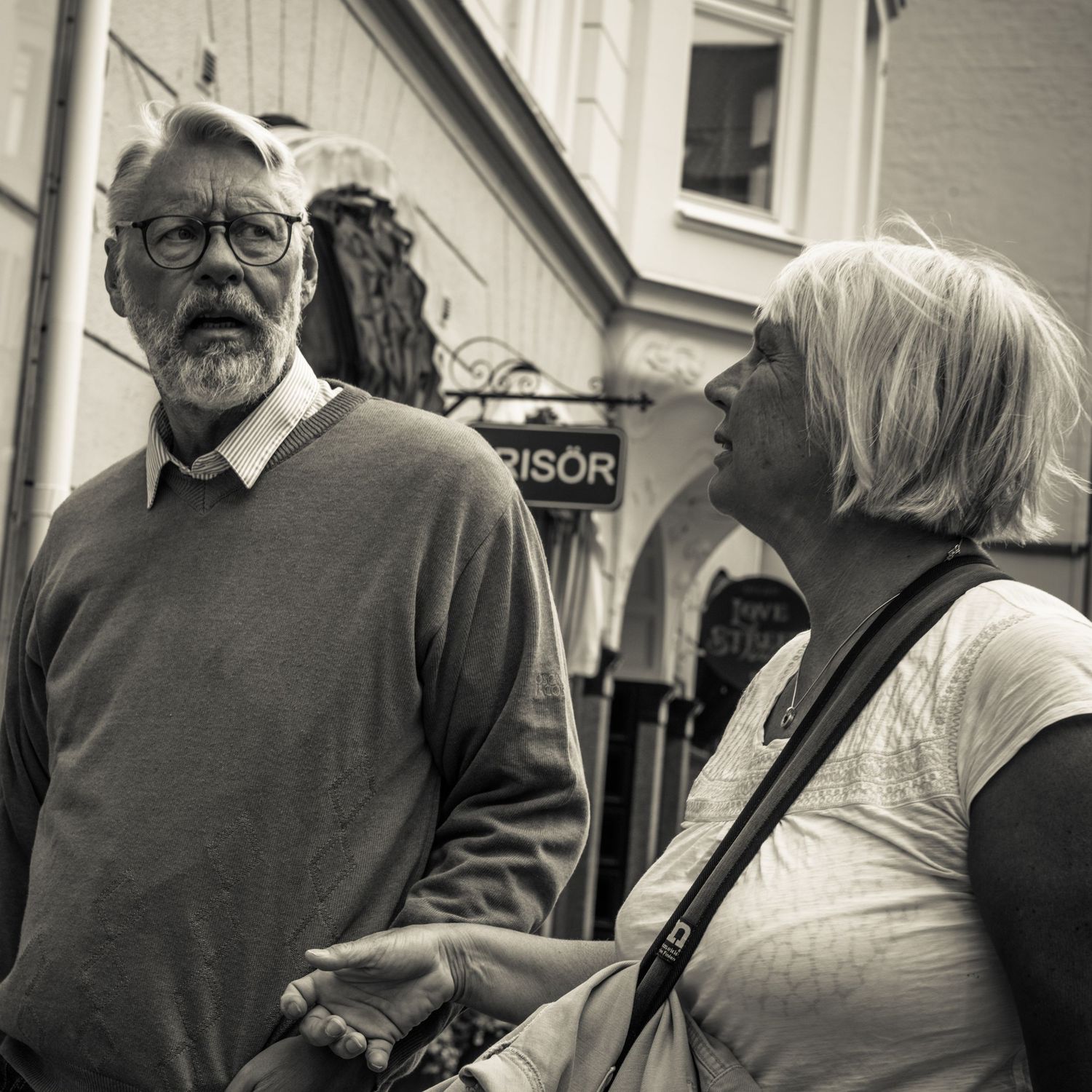 Elderly bearded man with glasses and woman with blonde hair stand together outdoors, black and white.