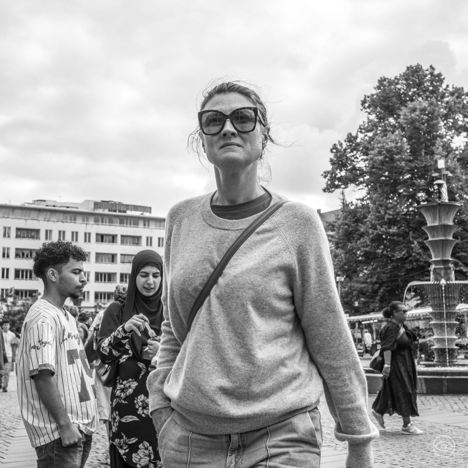 Woman in sunglasses and grey sweater walks through a busy city square in black and white street photography.