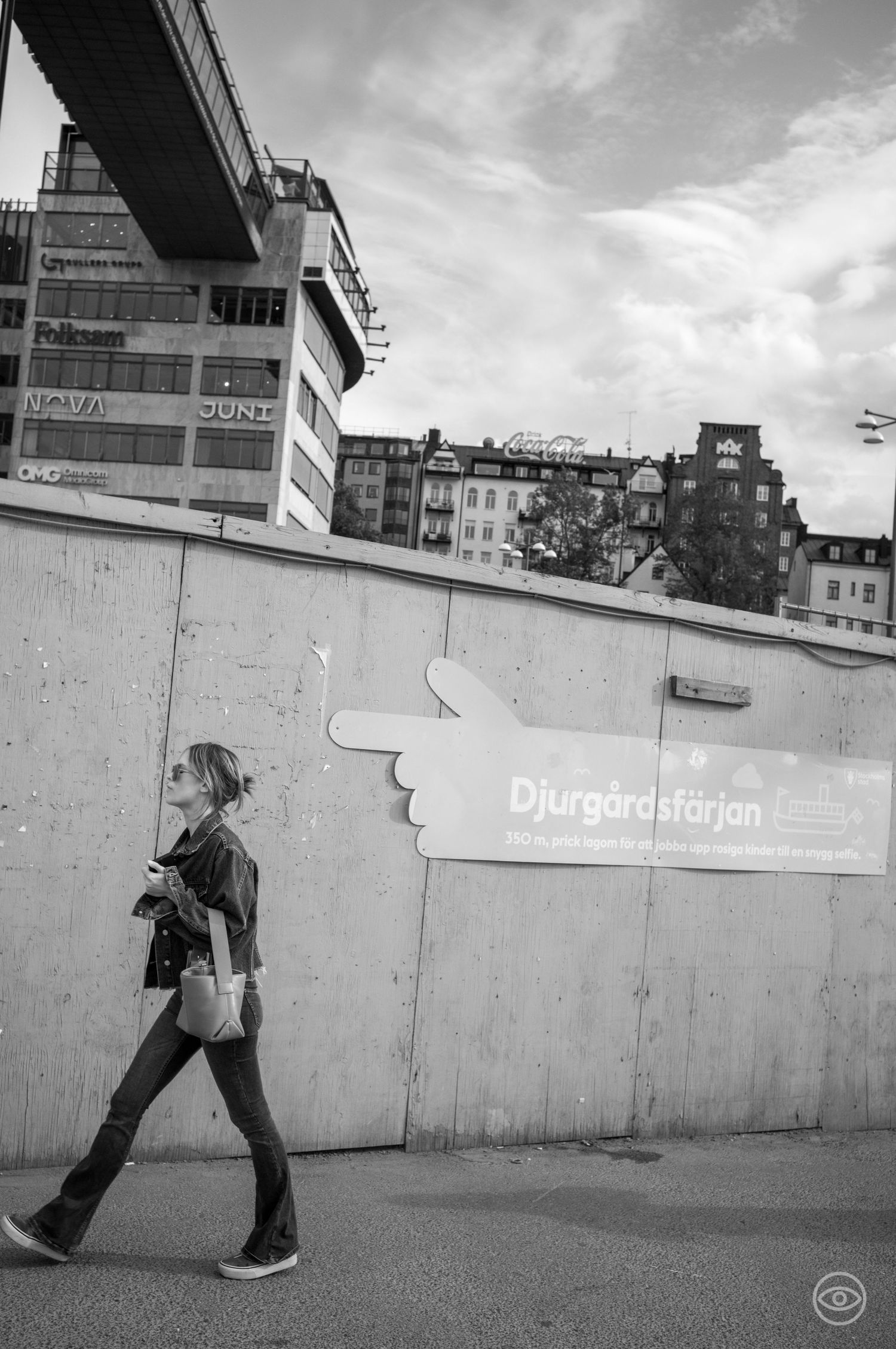 Woman walks past a Djurgardsfärjan sign on a concrete wall in black and white street photography.