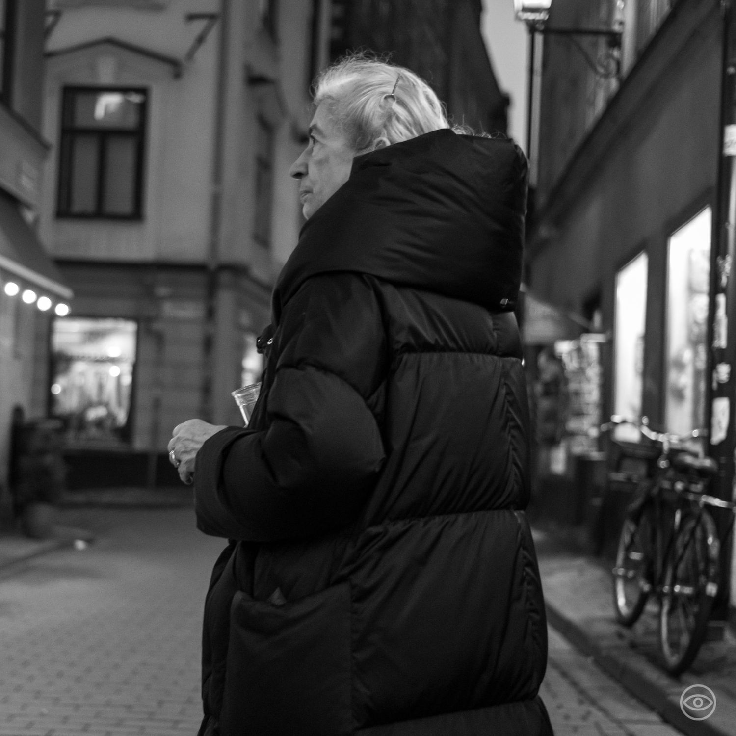 An elderly person in a large black puffer jacket walking along a city shopping street at night, black and white photography.