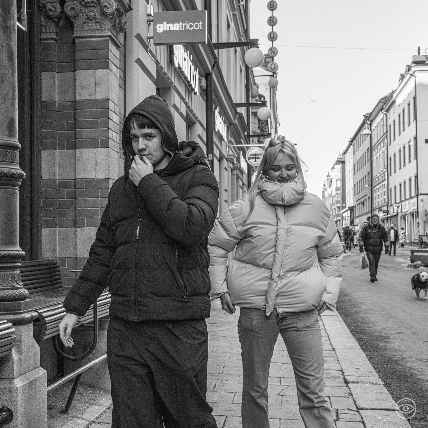 Two young people in puffer jackets walking down a city sidewalk past shops in black and white.