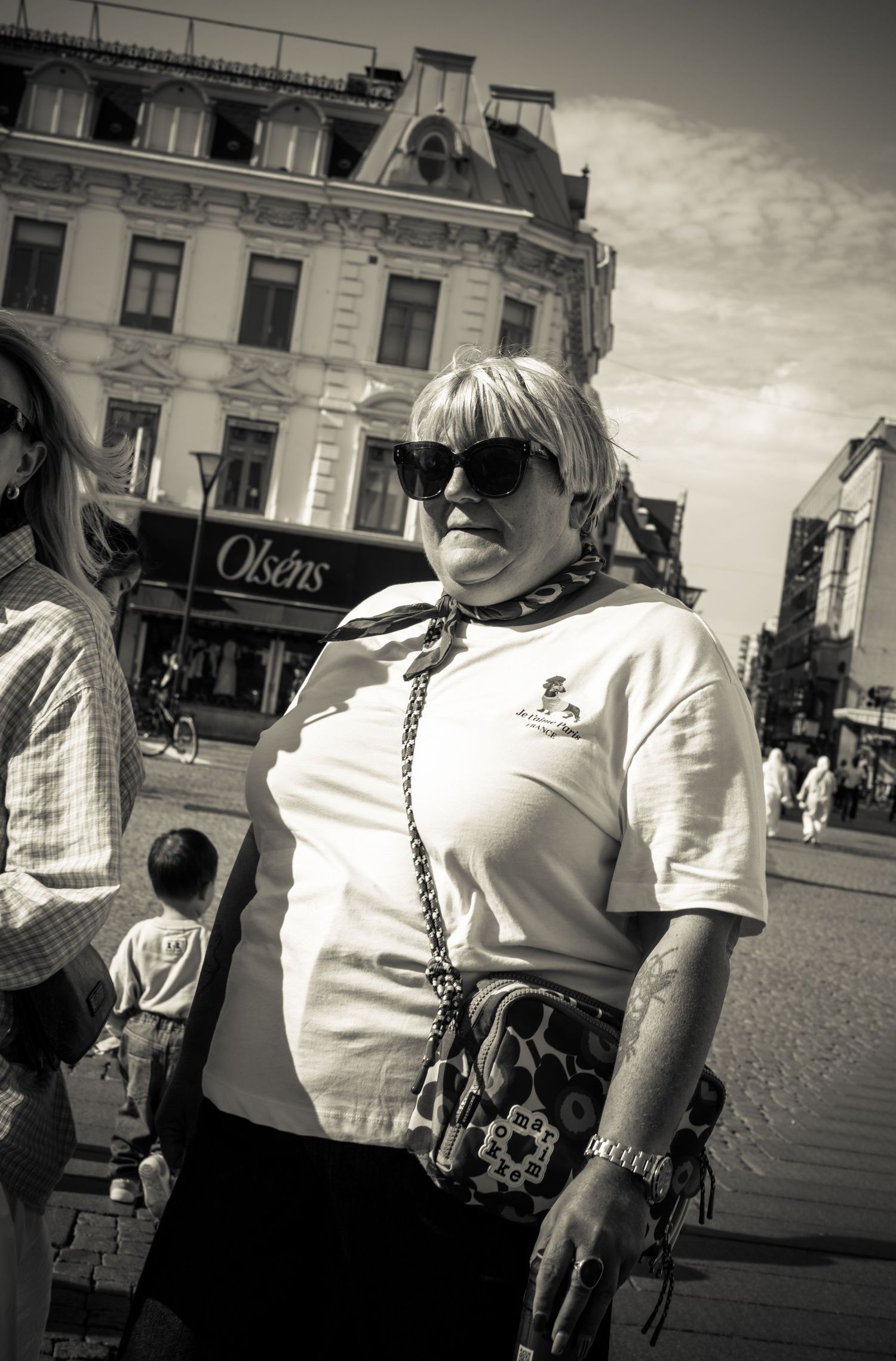 Older blonde woman in sunglasses and white t-shirt stands in city square, black and white.