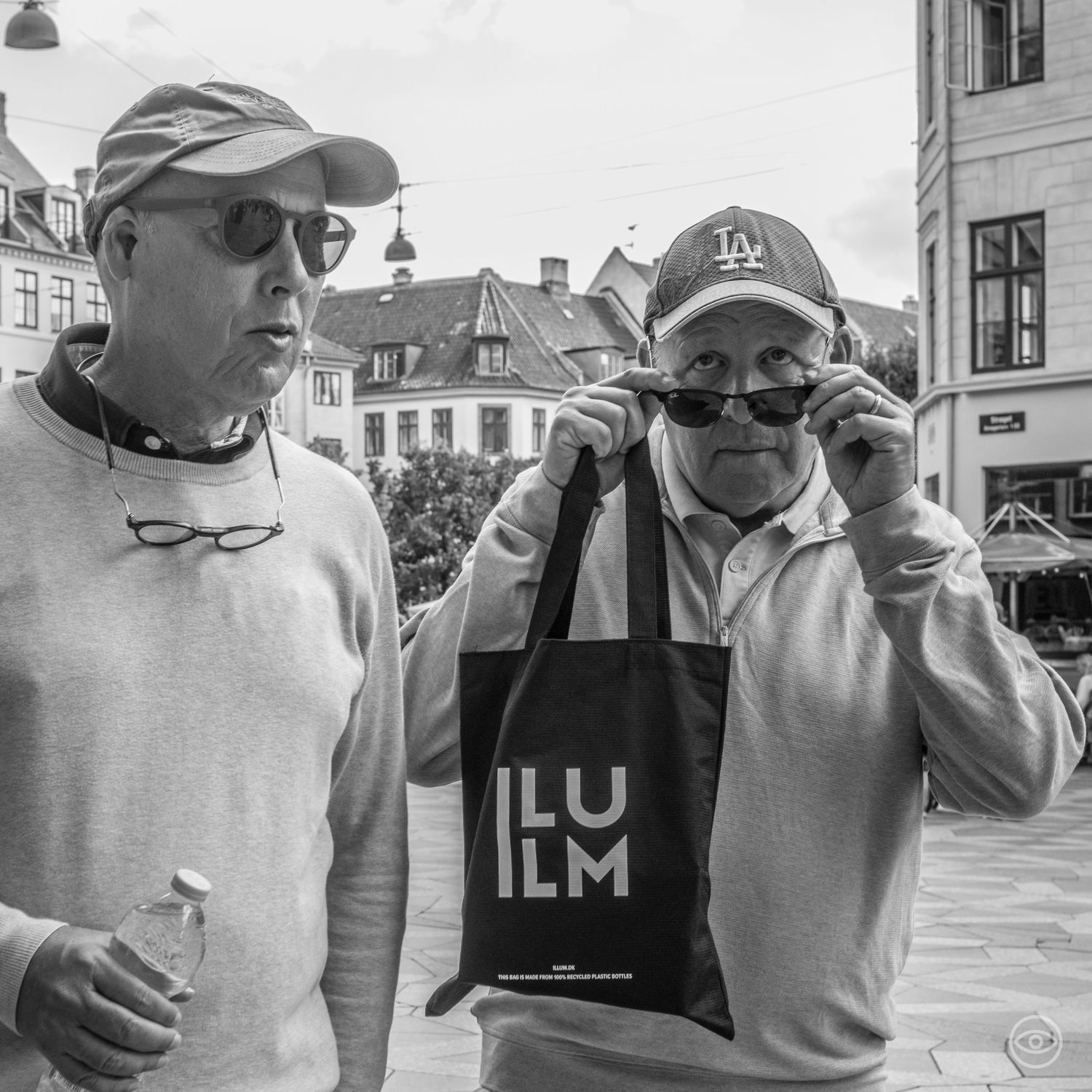 Two older men wearing caps and sunglasses holding an ILU ILM tote bag in a European town square, black and white photo.