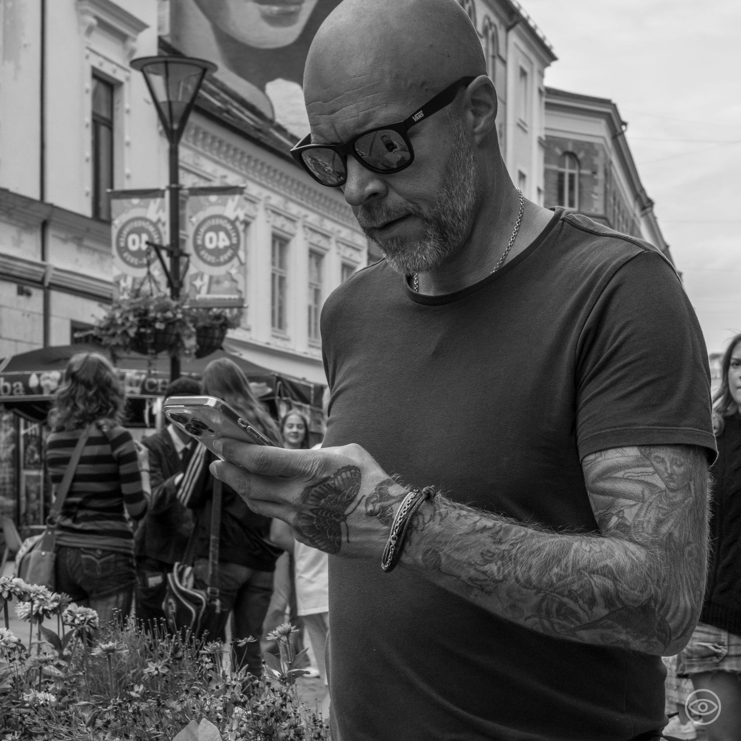 Tattooed man with glasses looking at his phone on a busy urban street in black and white.