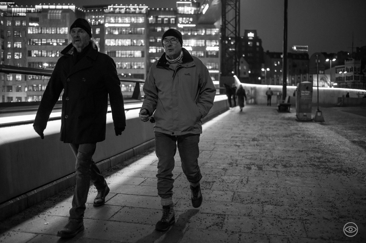 Two men walking side by side on a city bridge at night with illuminated buildings in the background, black and white photo.