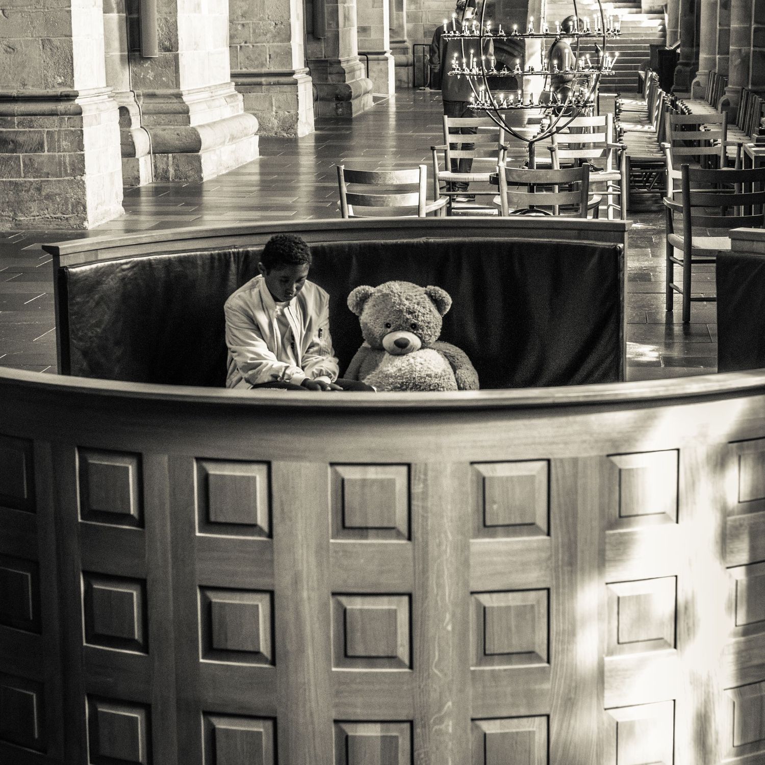 Man sits beside a large teddy bear inside a wooden pulpit in a black and white church interior.
