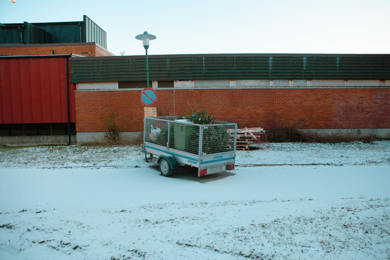Small utility trailer with a Christmas tree parked on snow-covered ground near brick building.