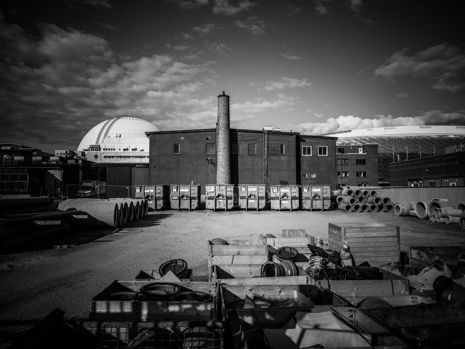 Black and white industrial yard with dome structure, chimney, brick building, and scattered machinery.