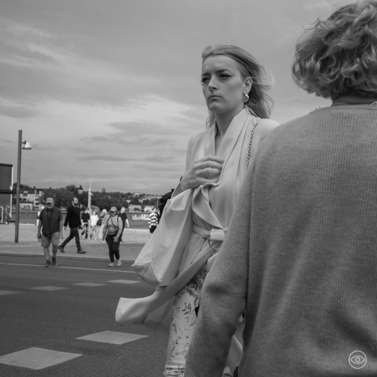 Blonde woman carrying a tote bag stands on a waterfront street in black and white street photography.