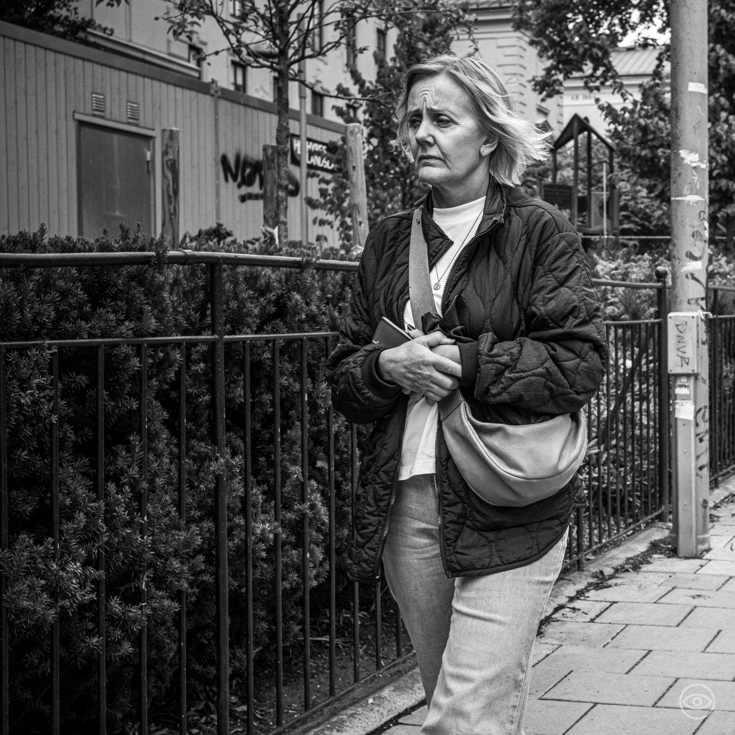 Middle-aged blonde woman with a shoulder bag leaning against an iron fence on a city street.