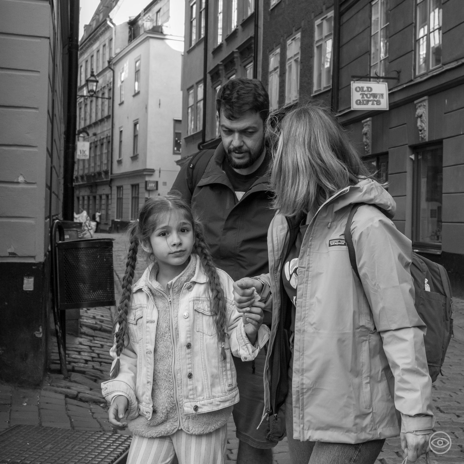 A man with two young girls converses on a cobblestone European street in a black and white photo.
