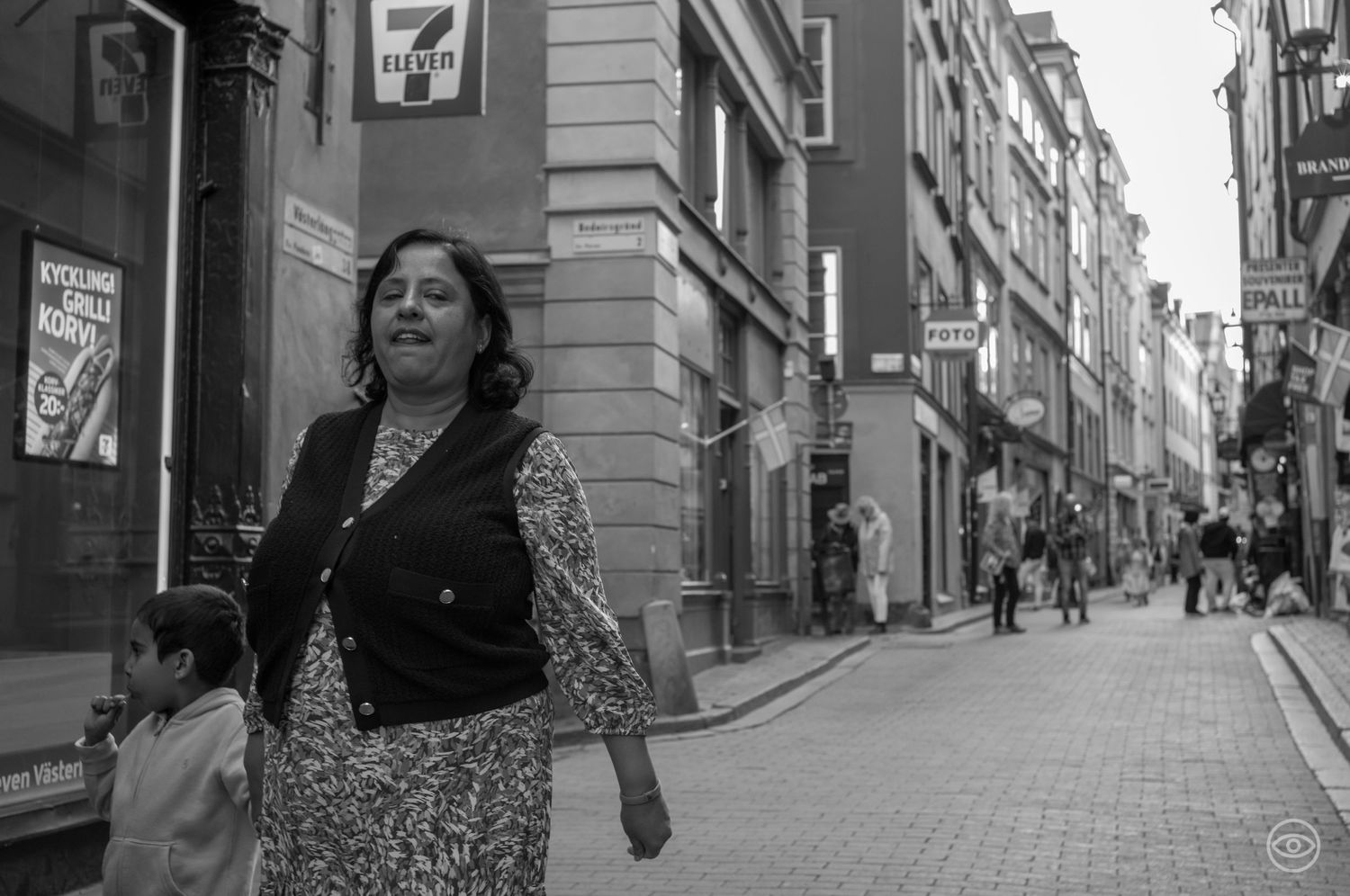 Woman in floral dress and black vest walking on a city street near a 7-Eleven sign.