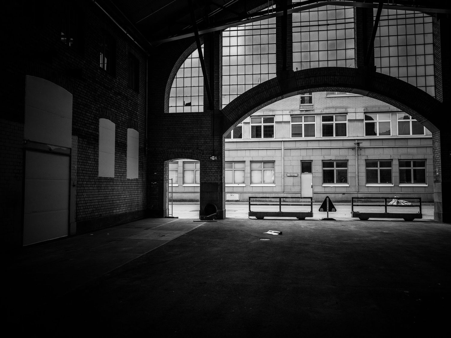 Lone figure sits on a bench inside a dark abandoned industrial hall with arched windows.