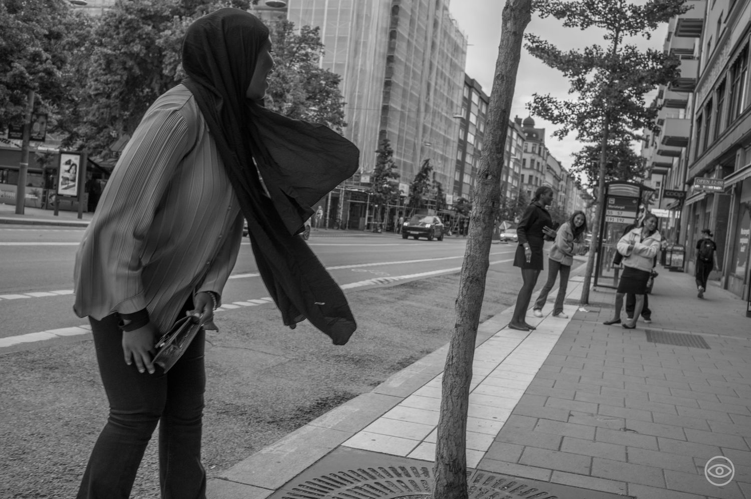 Woman wearing a hijab walking along an urban sidewalk with trees and city street in background.