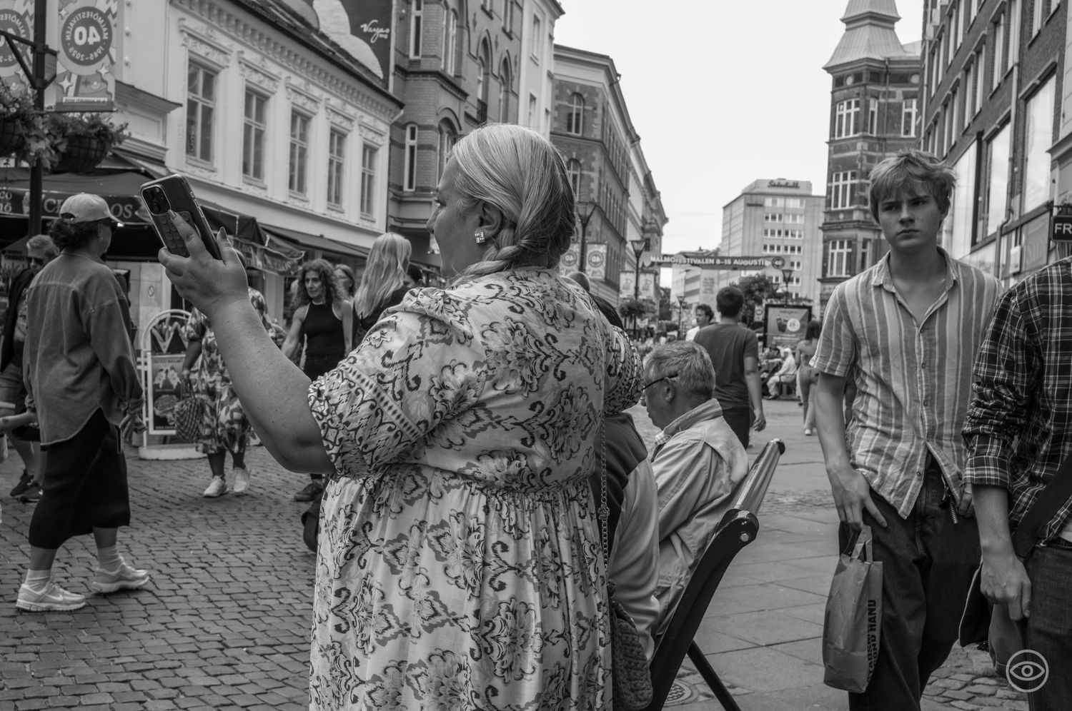 Older woman in floral dress taking a photo on a busy pedestrian shopping street in black and white.