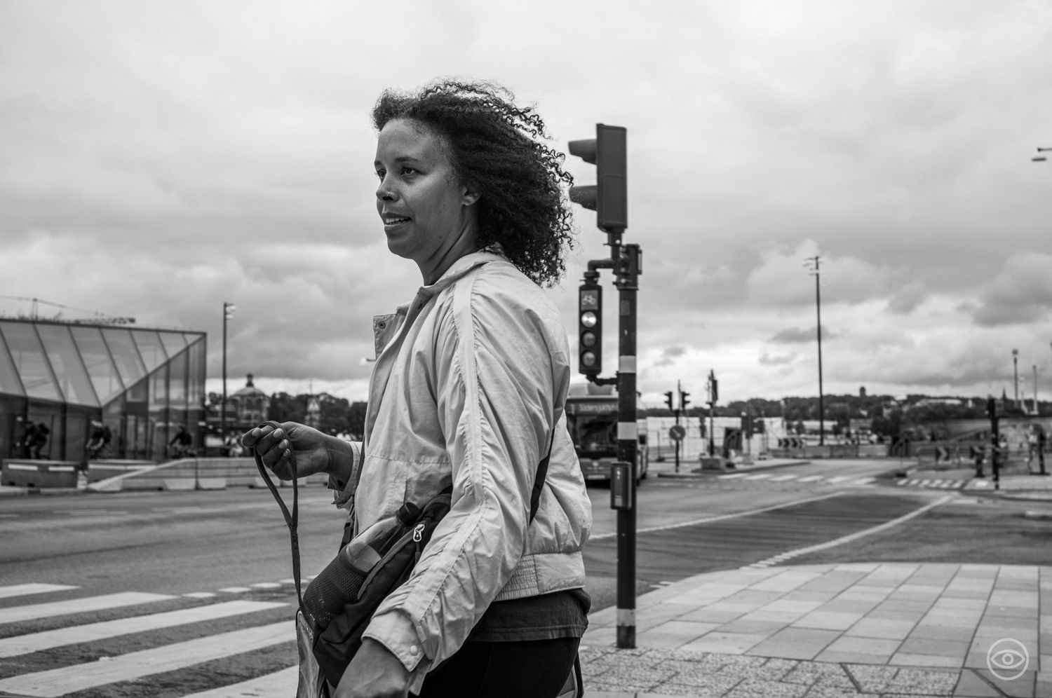 Woman with curly hair standing at a crosswalk on a cloudy day, black and white street photo.