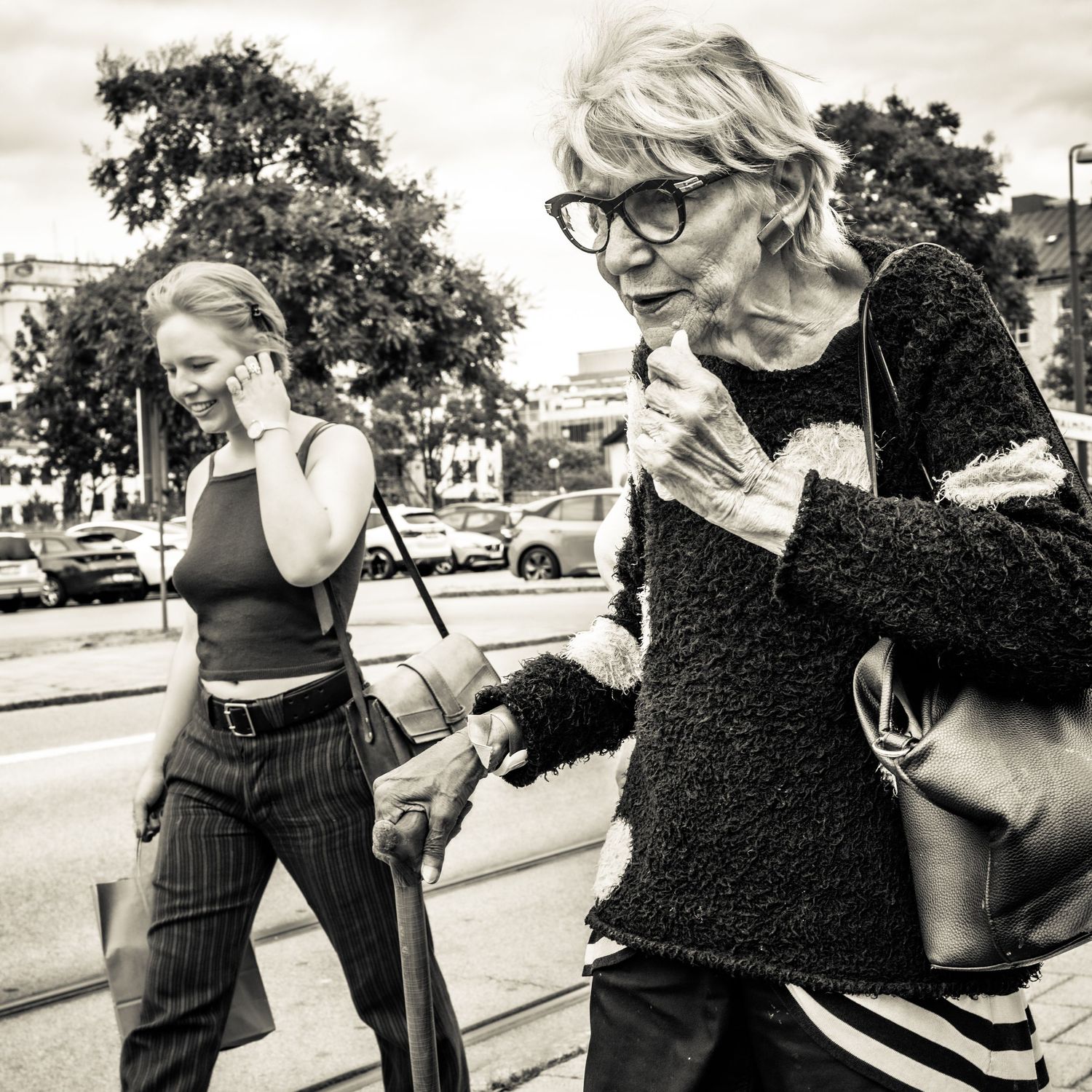 Black and white street photo of two women walking, one elderly with a cane and glasses, another younger on phone.