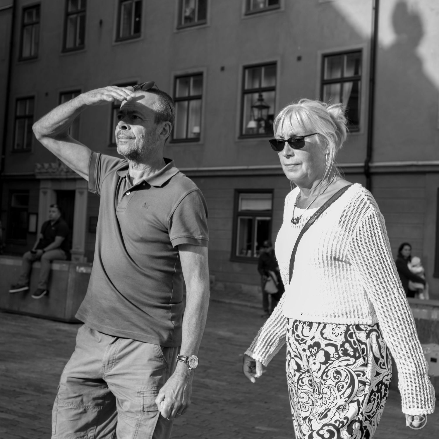 Elderly couple walks in a sunny city square, man shielding his eyes from the sun in black and white.