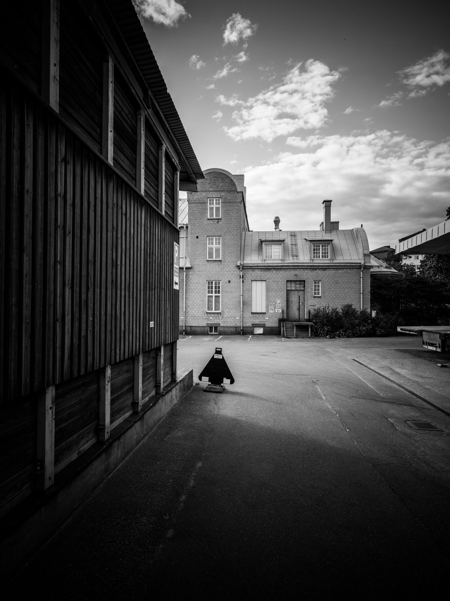 Solitary figure sits on ground beside a tall wooden wall near an old building.