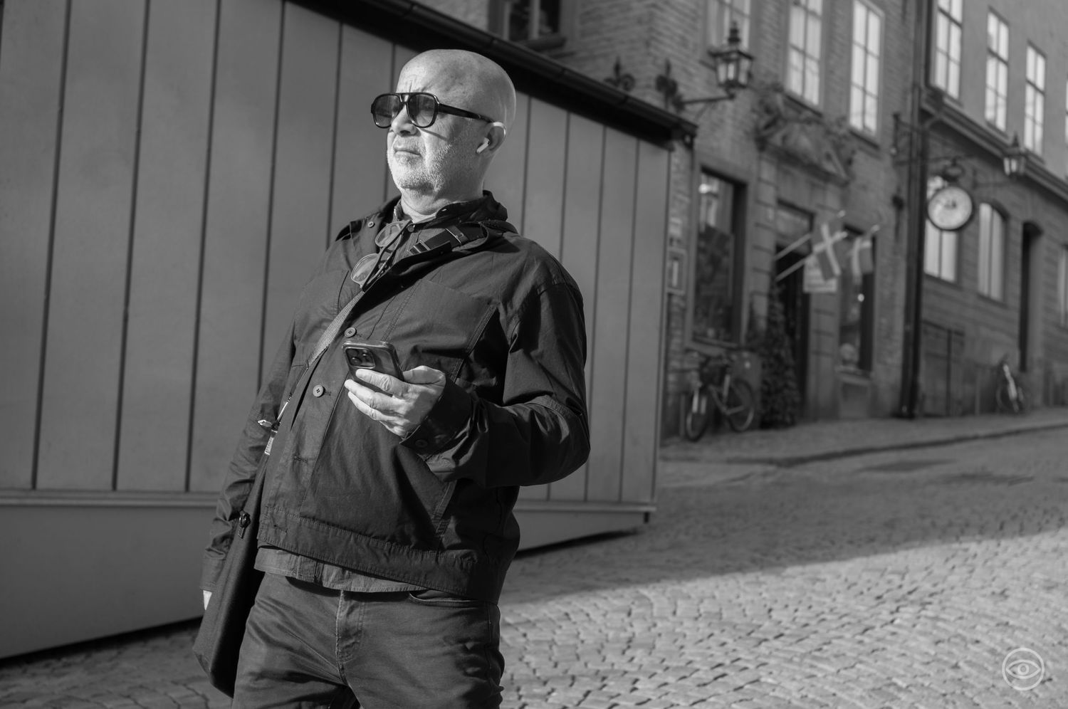 Bald man wearing sunglasses and black jacket holding a camera on a cobblestone street in black and white.