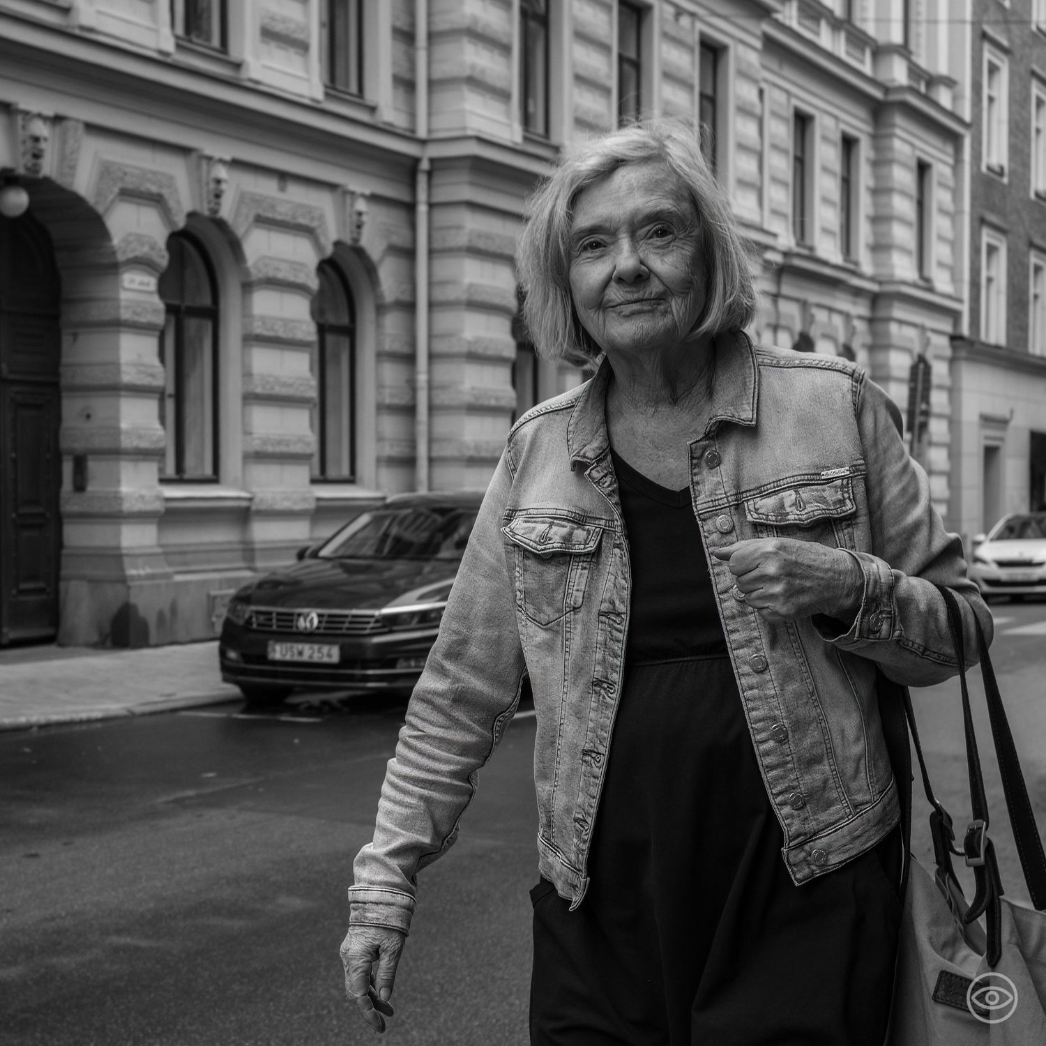 Elderly woman in a denim jacket walking on a city street, black and white street photography.