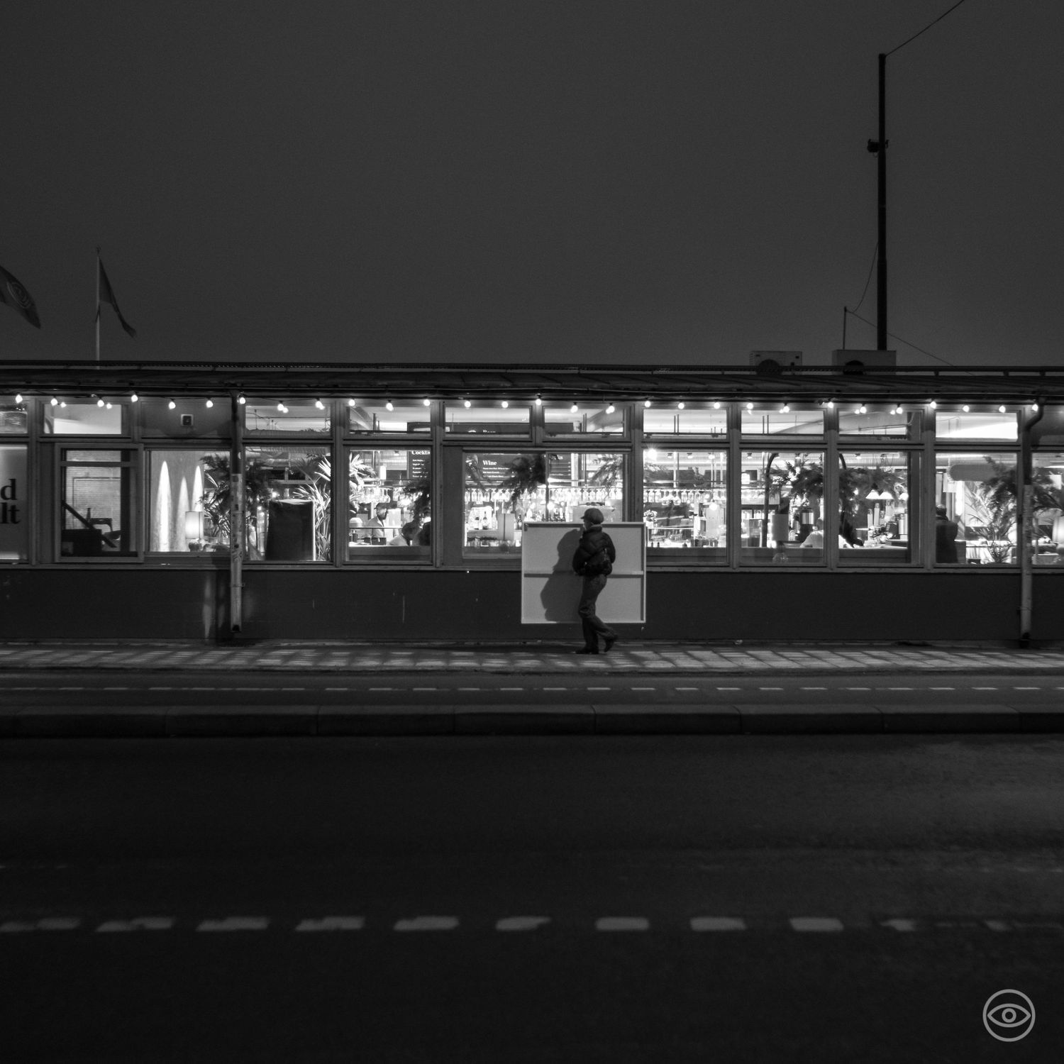 A lone figure sits at an illuminated bus stop or tram station at night on an empty urban street, black and white photo.