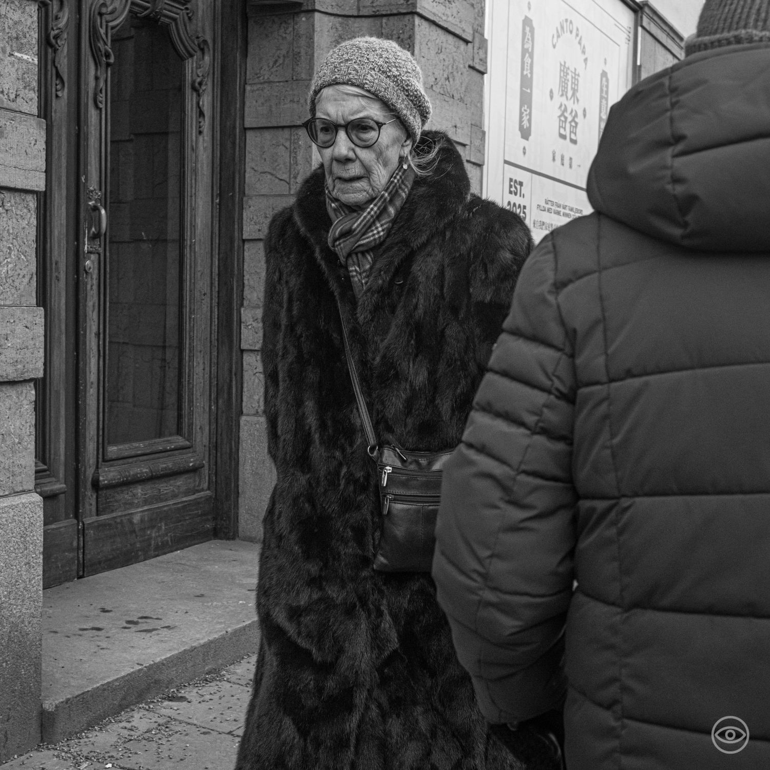 Elderly woman in a long fur coat and knit hat wearing glasses on a city street in black and white.