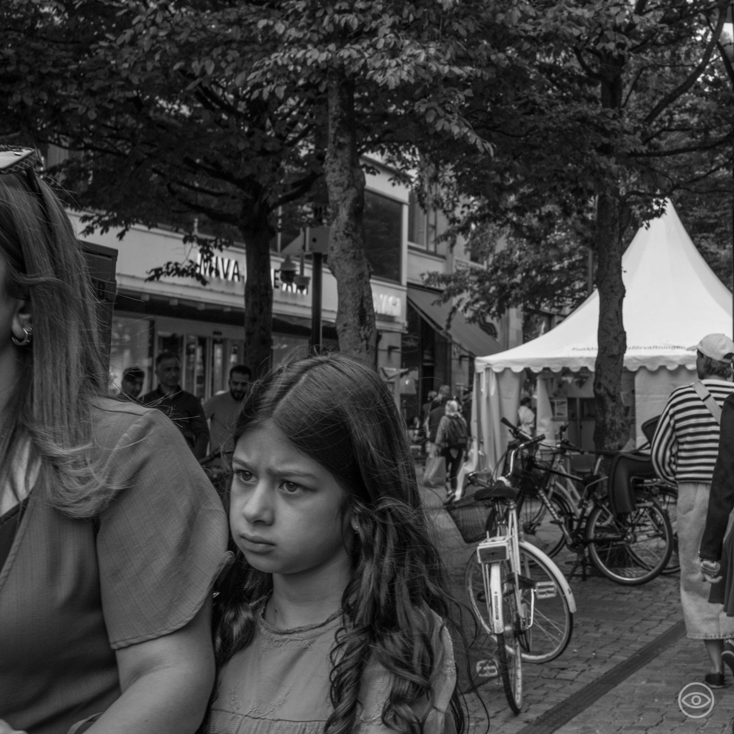 Young girl with a serious expression sits beside a woman at an outdoor city market with trees and tents.